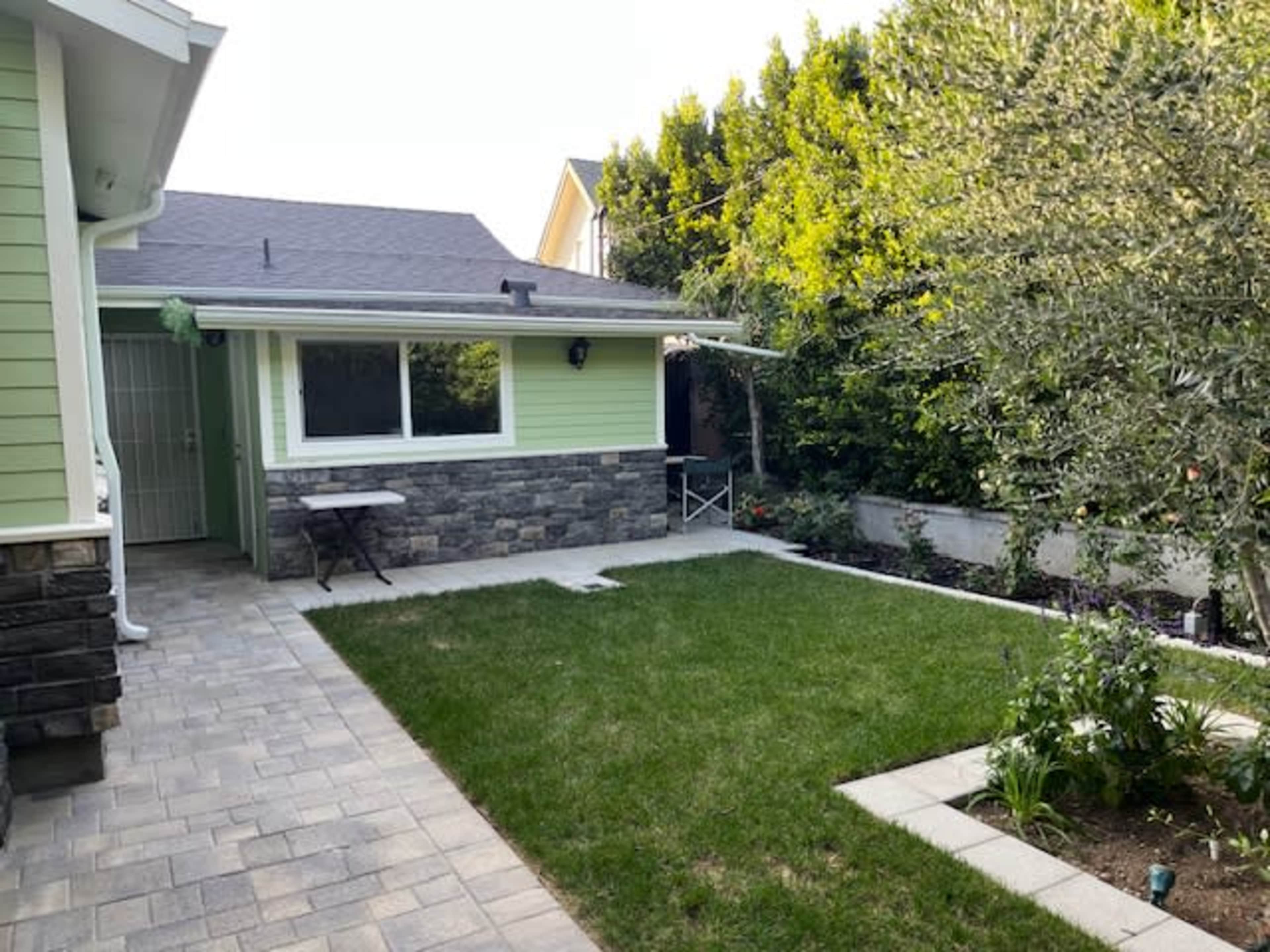 The image shows a well-maintained backyard featuring a green lawn, stone patio, and a house with light green siding.