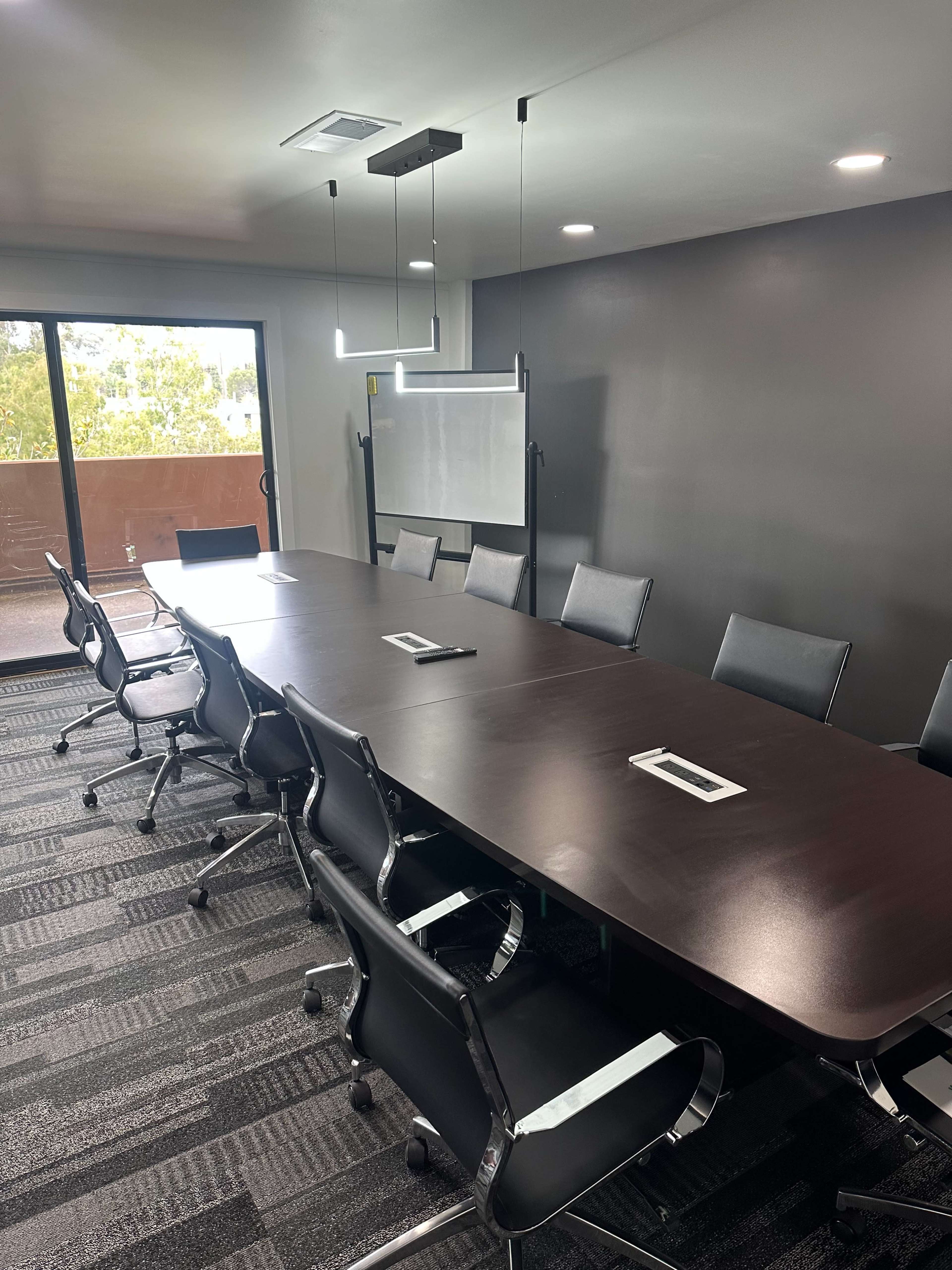 A large conference room features a long wooden table surrounded by black chairs, with a whiteboard on a stand and windows offering natural light.