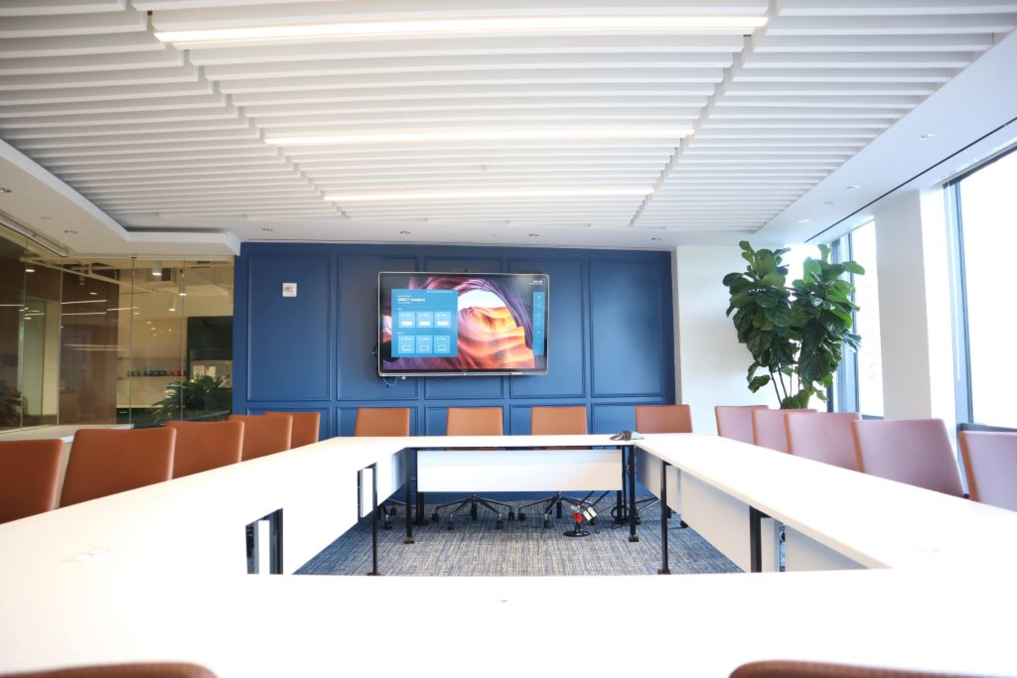A modern conference room features a large screen on the wall and a U-shaped layout of white tables surrounded by brown chairs.