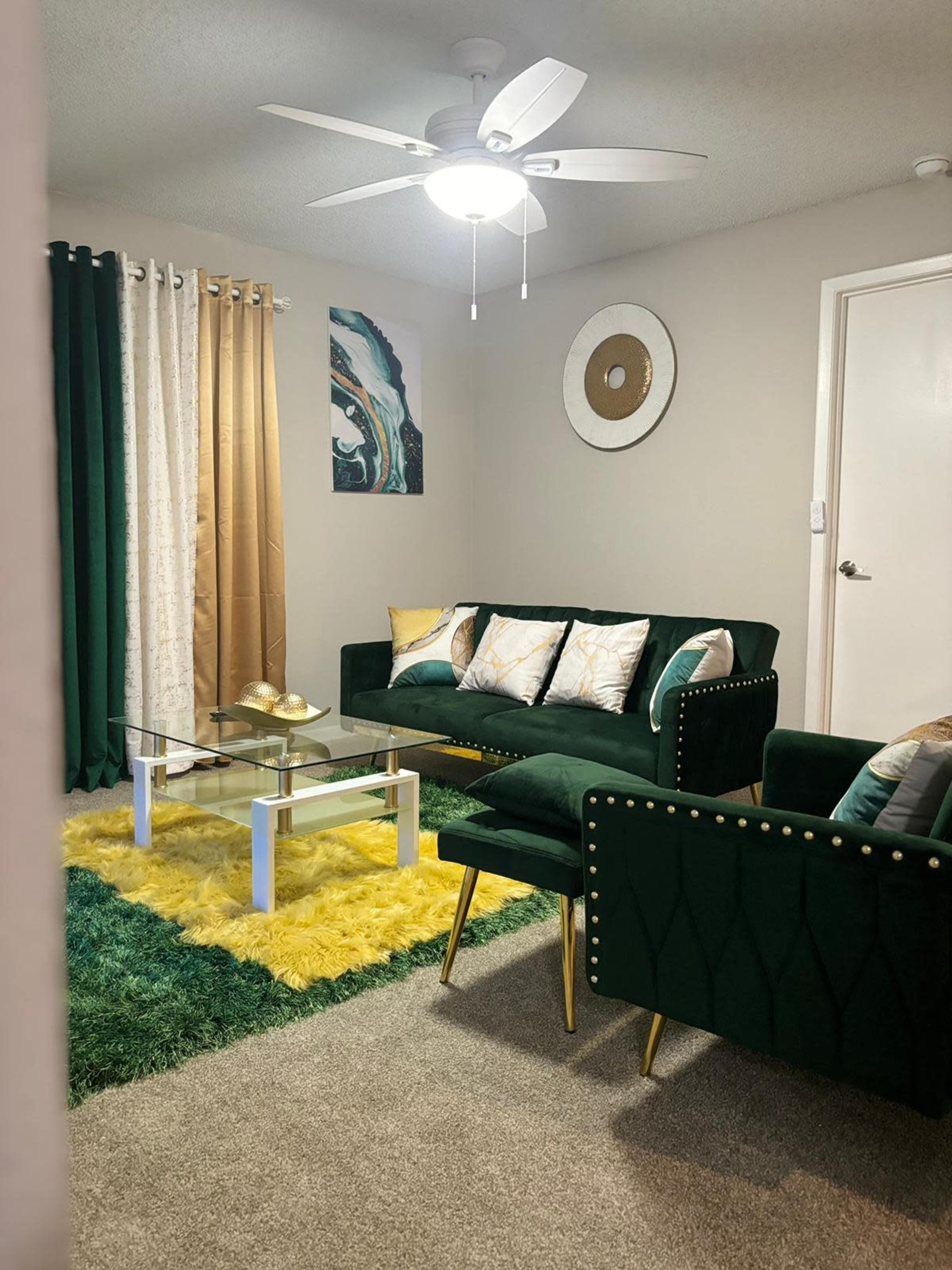 A modern living room furnished with a green sofa, a glass coffee table, and a yellow shag rug, with decorative curtains and wall art in the background.