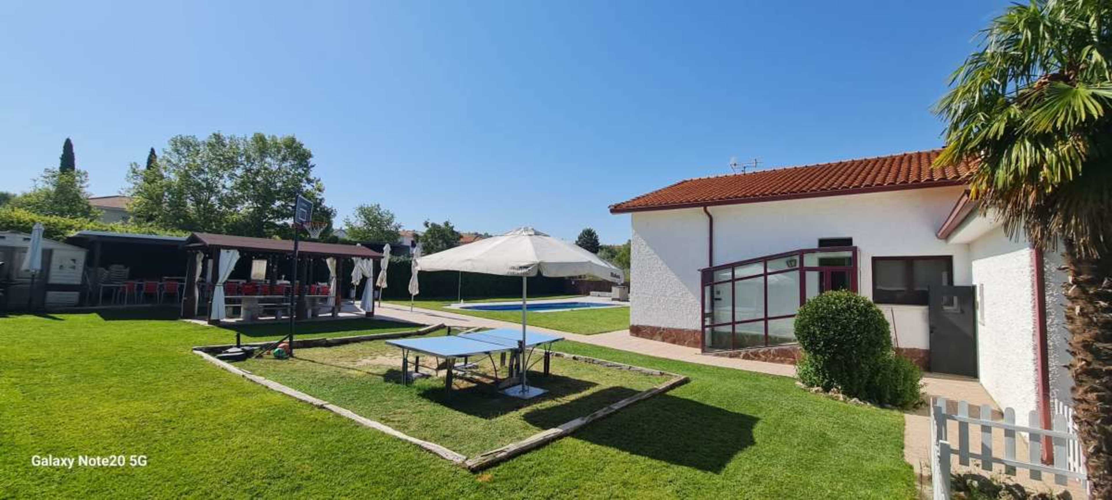 The image shows a well-maintained backyard featuring a green lawn, a table tennis setup, a large umbrella, and a covered seating area beside a house.