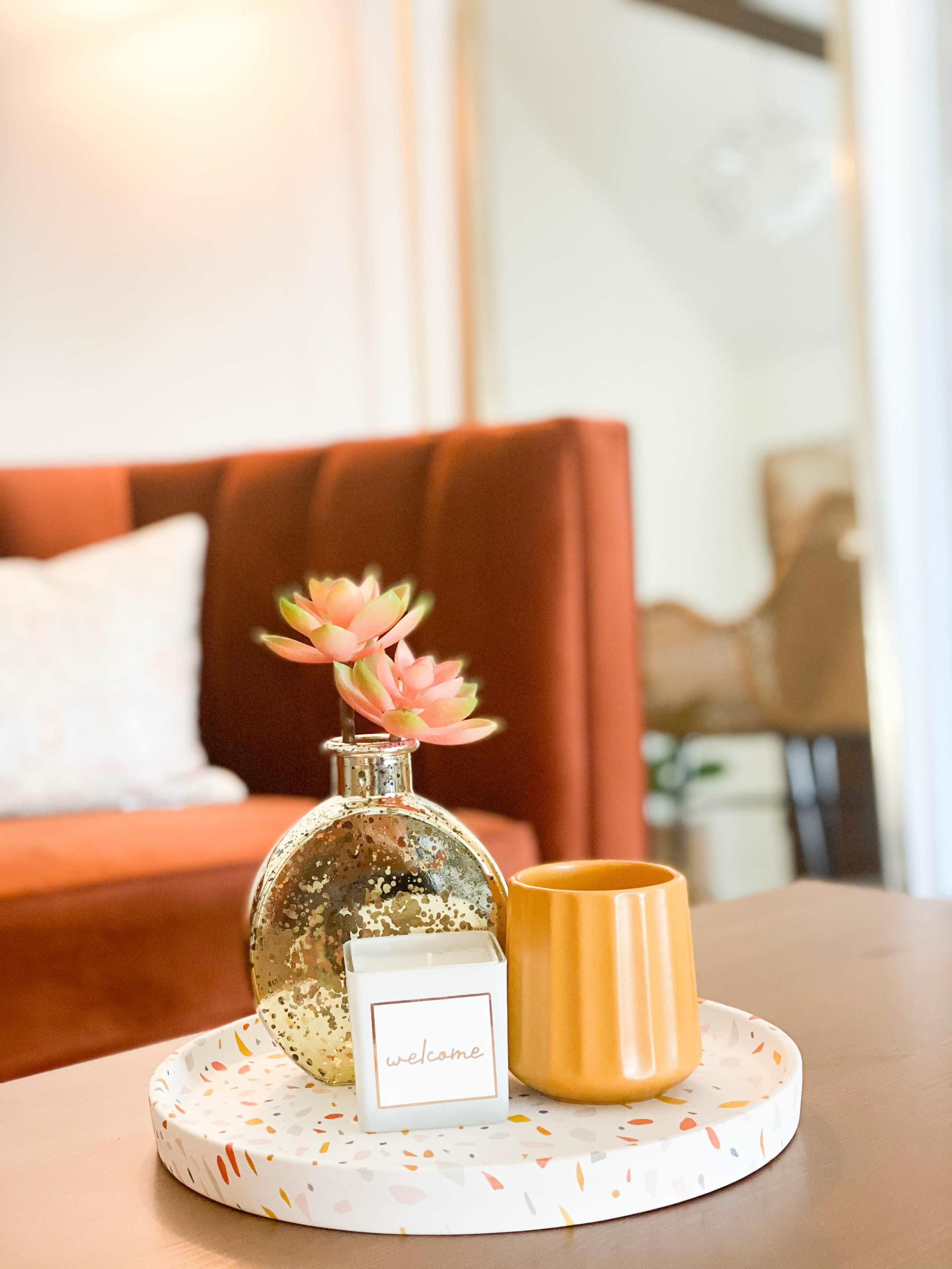 A decorative tray on a coffee table holds a flower vase, a small orange cup, and a welcome sign, set against a softly lit living room.