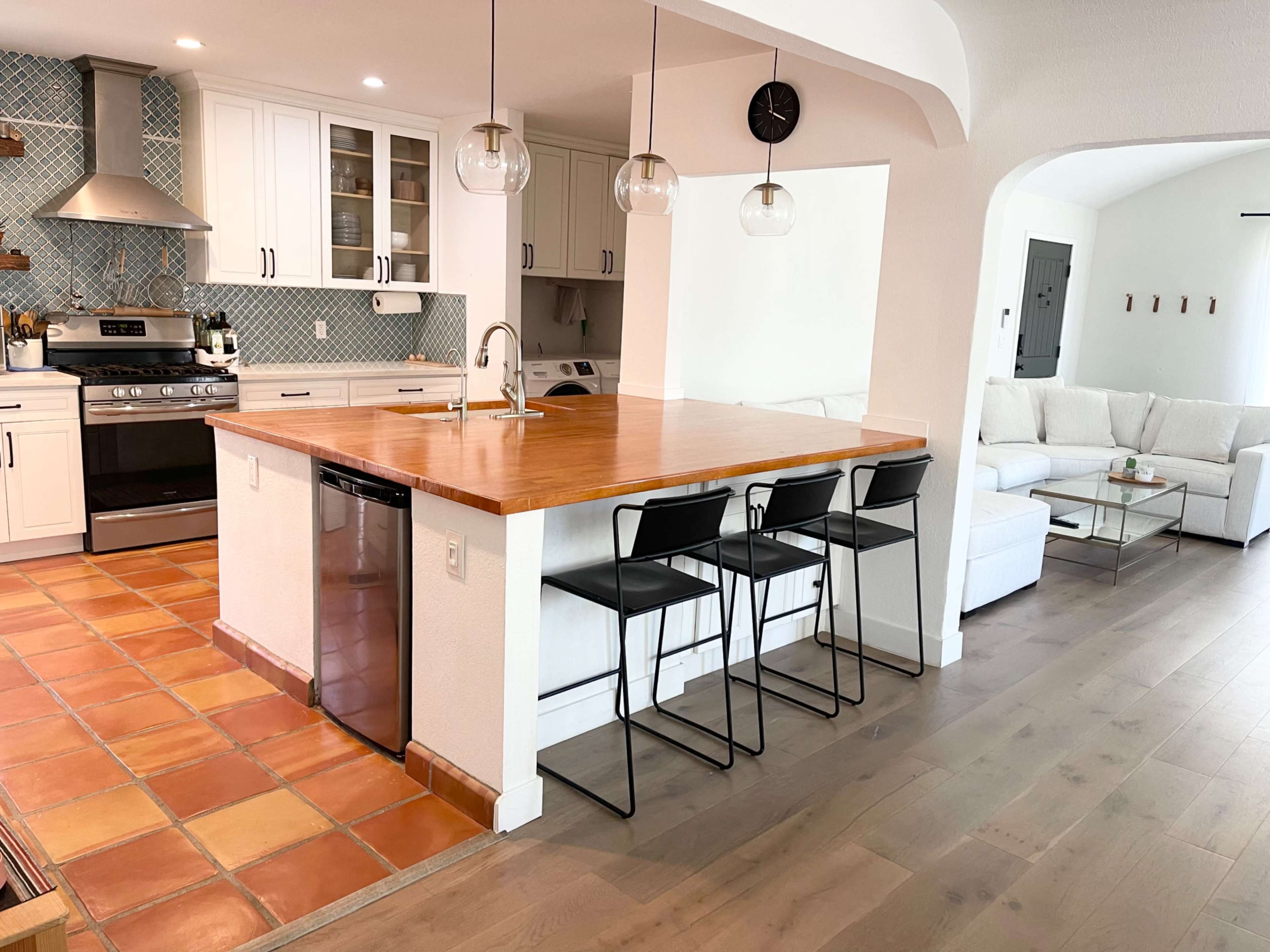 A modern kitchen with a wooden island, three black stools, and a cozy living area in the background featuring a white sofa and coffee table.