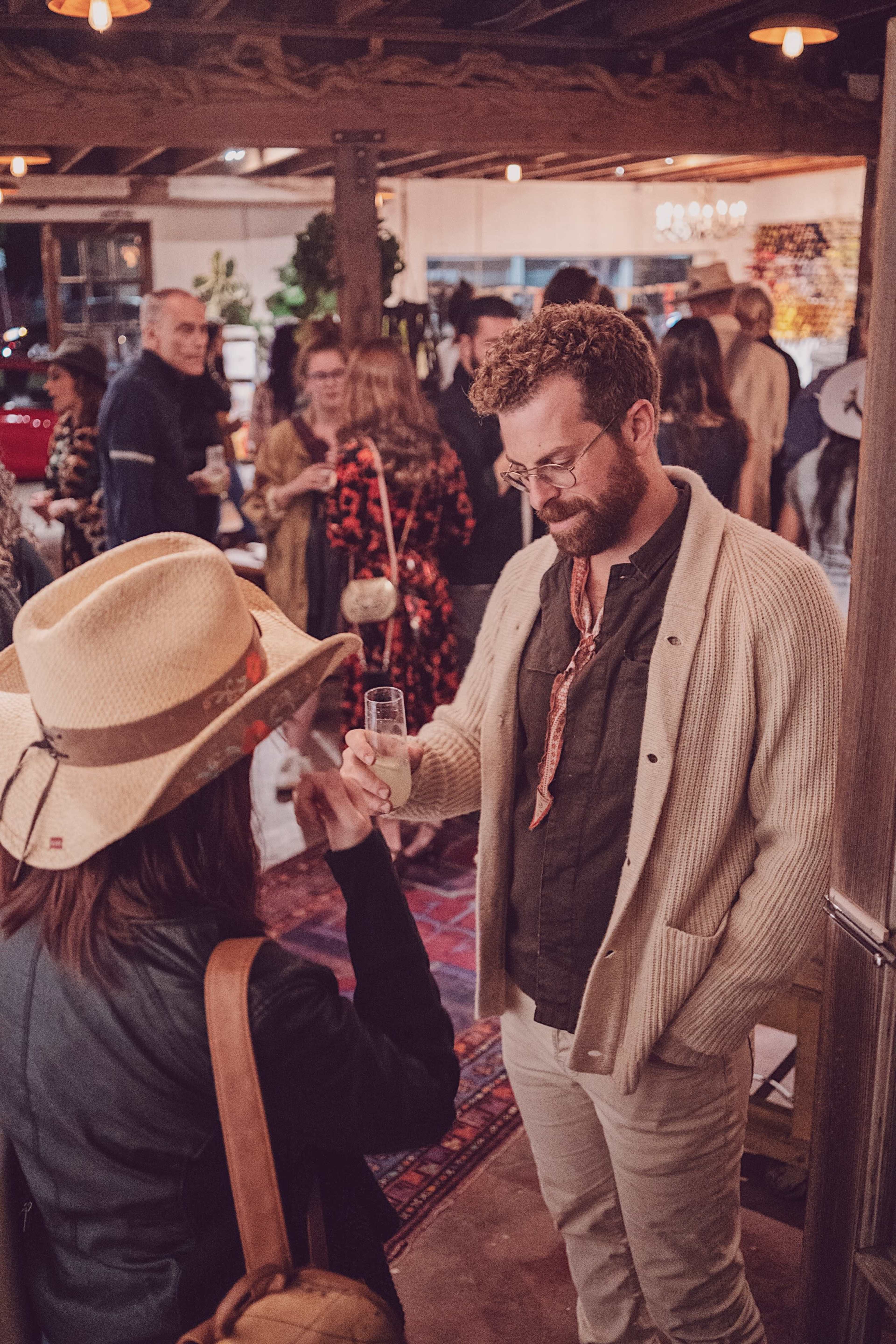 A man in a sweater and glasses holds a drink while conversing with a woman wearing a wide-brimmed hat at a crowded indoor event.