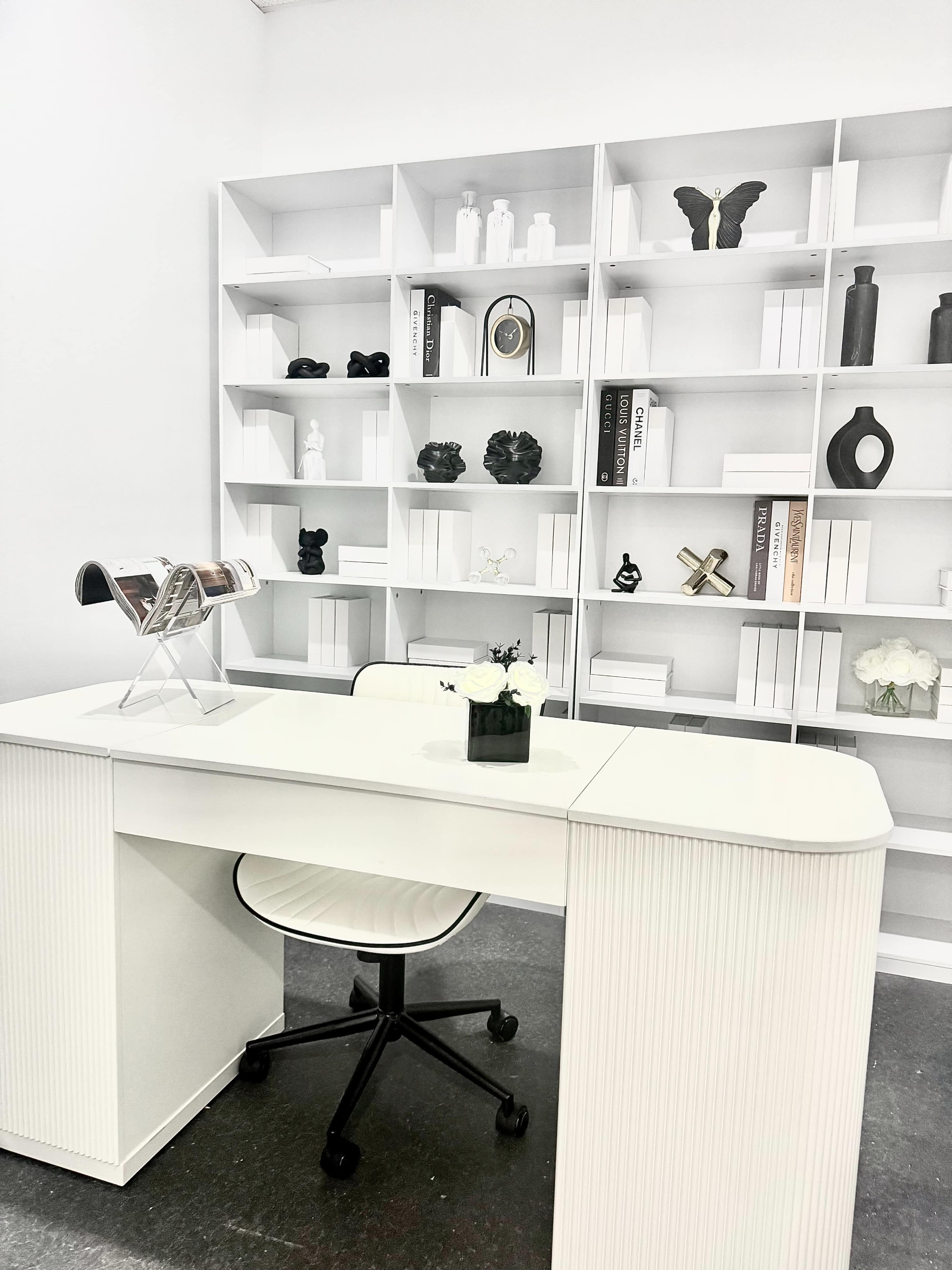 The image shows a modern office space with a white desk and a shelf filled with decorative items and books.