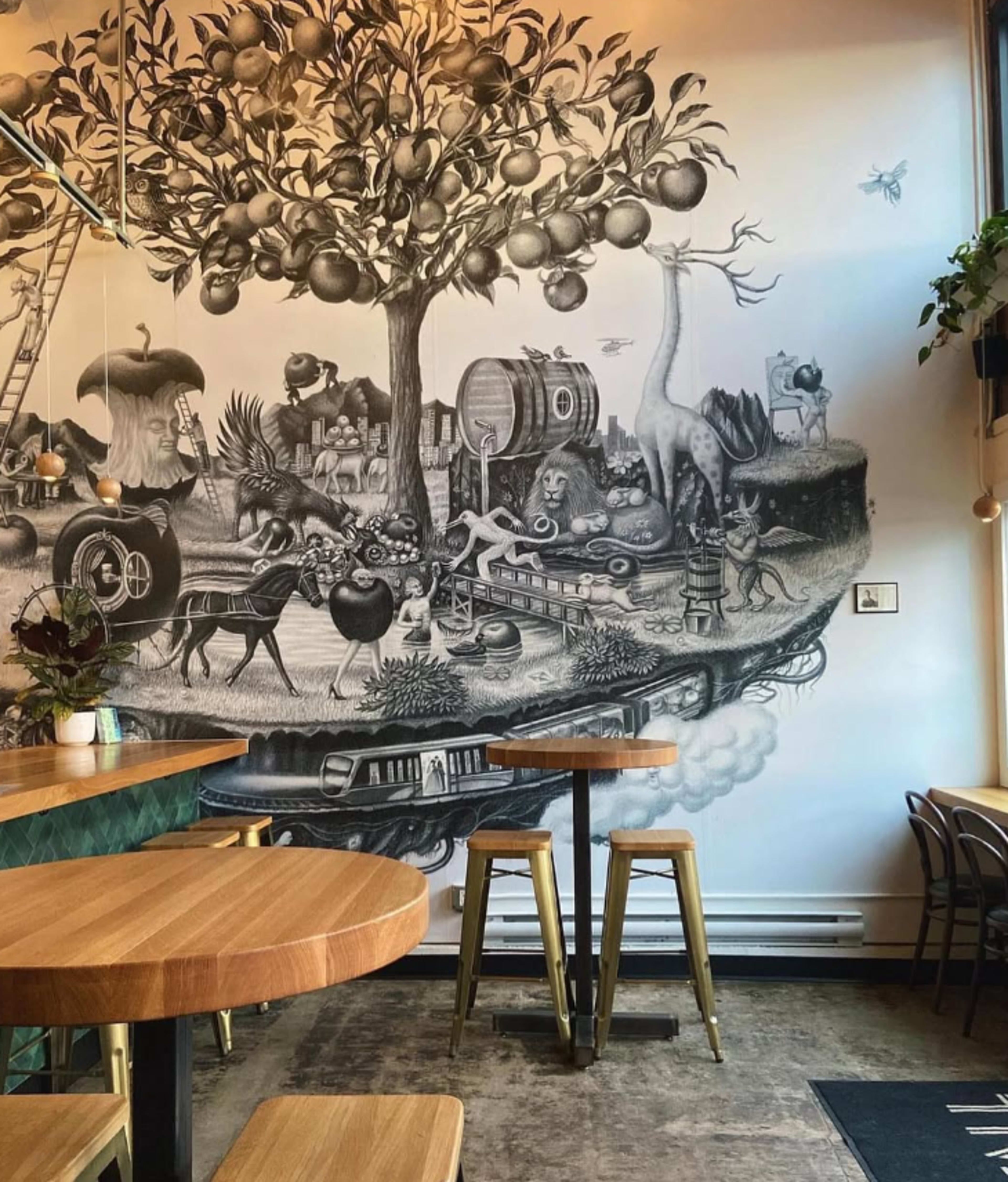 The image features a detailed mural depicting a fantastical scene with various animals and objects integrated into a surreal landscape, set against the wall of a cafe with wooden tables and chairs.