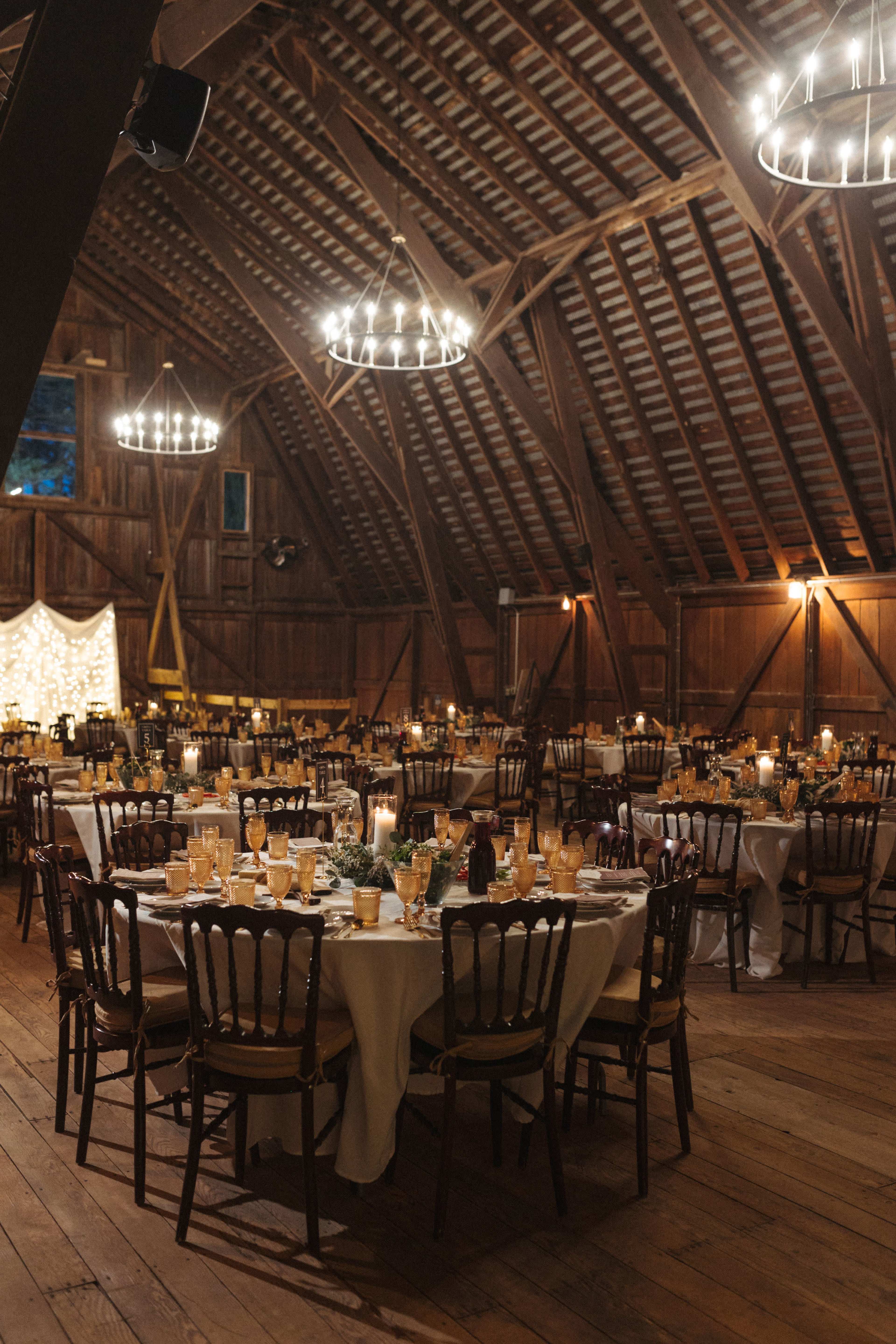 A rustic barn interior set up for a banquet, featuring several round tables covered with white tablecloths and surrounded by wooden chairs, illuminated by chandelier lights.
