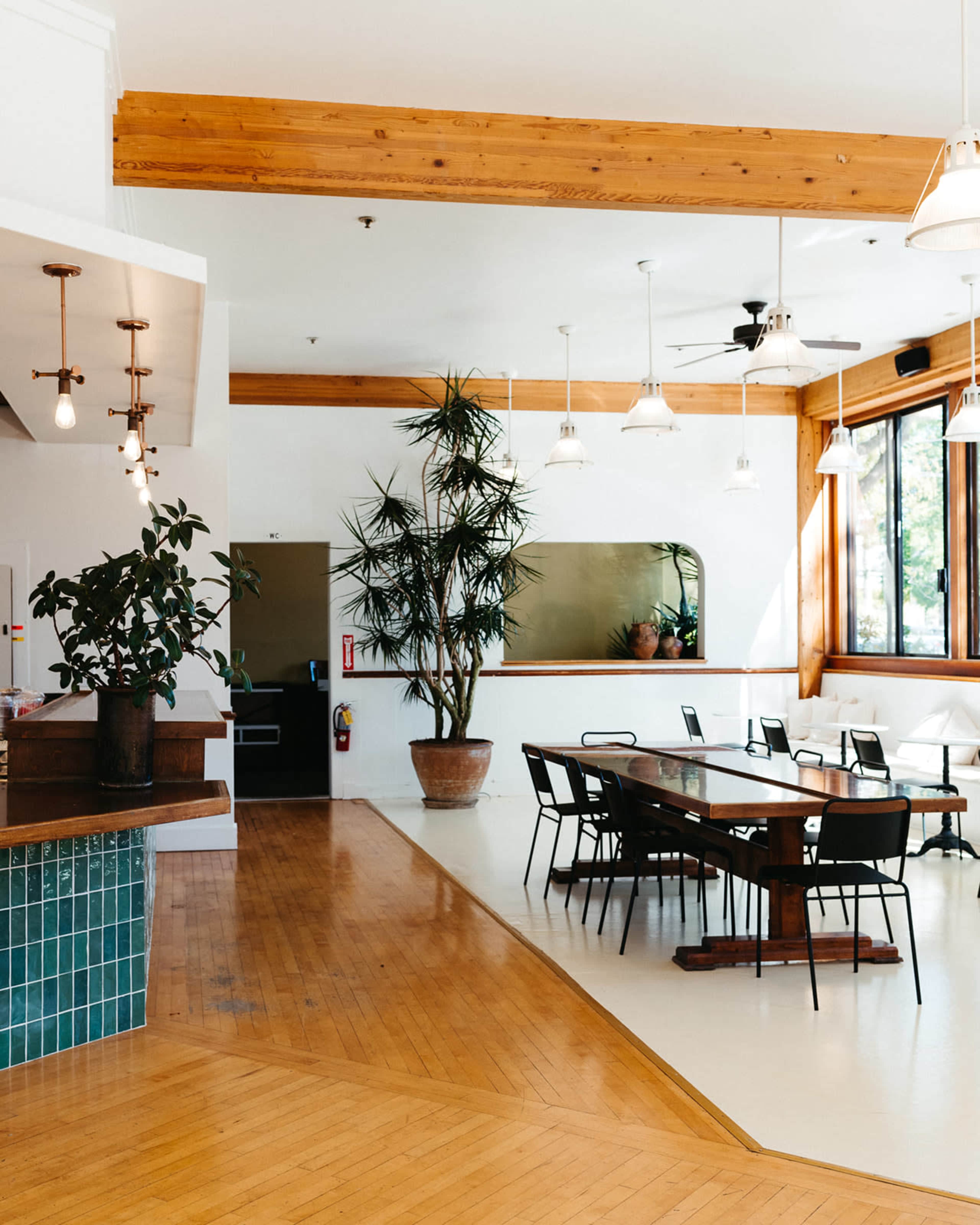 A bright, spacious dining area featuring a wooden table surrounded by black chairs, framed by large windows and indoor plants.