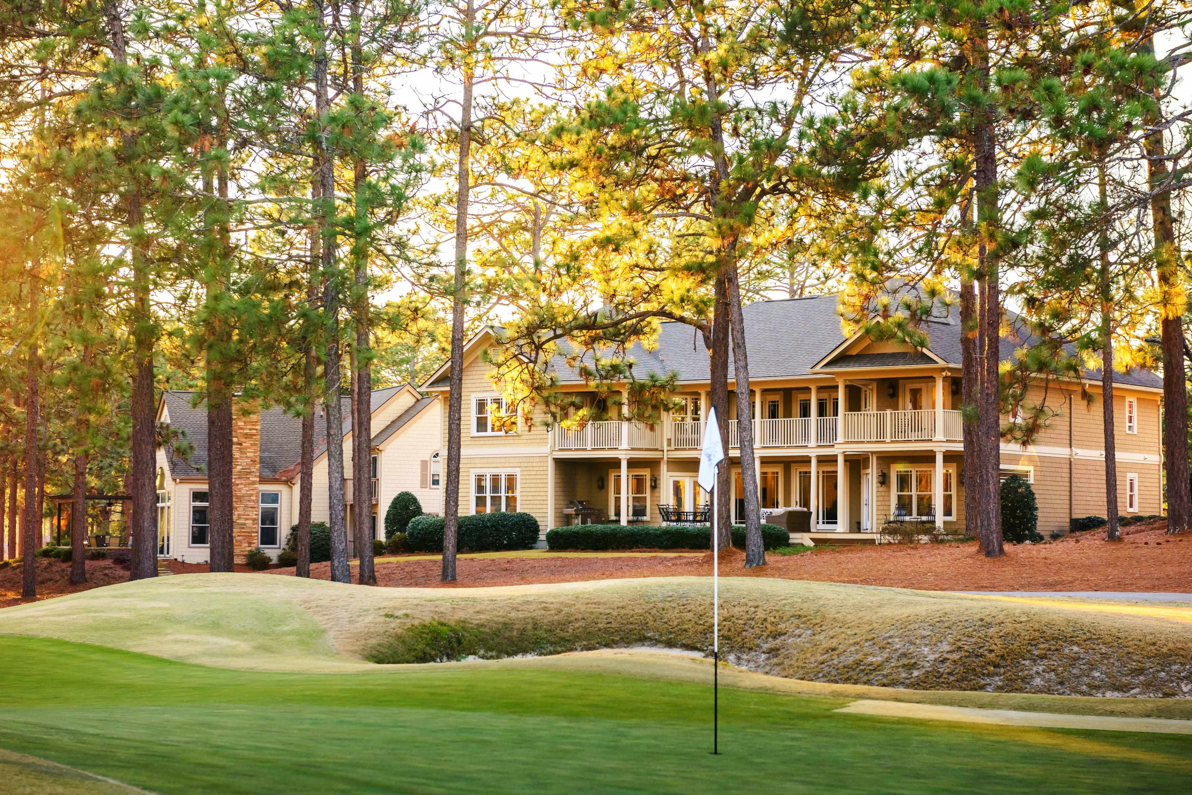 A golf course with a putting green in the foreground and several wooden houses nestled among tall pine trees in the background.