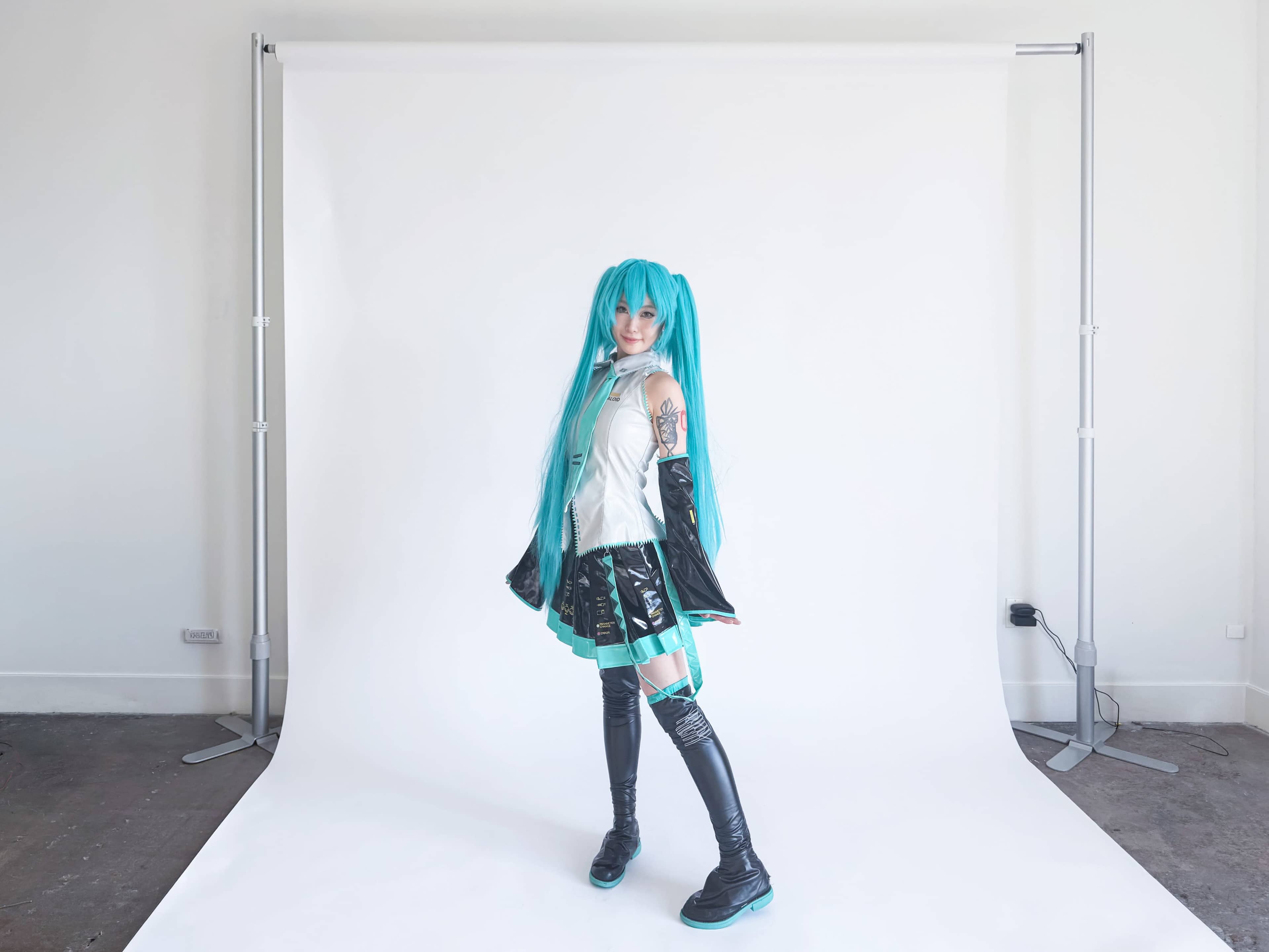 A person with turquoise hair and a costume stands in front of a plain white backdrop.