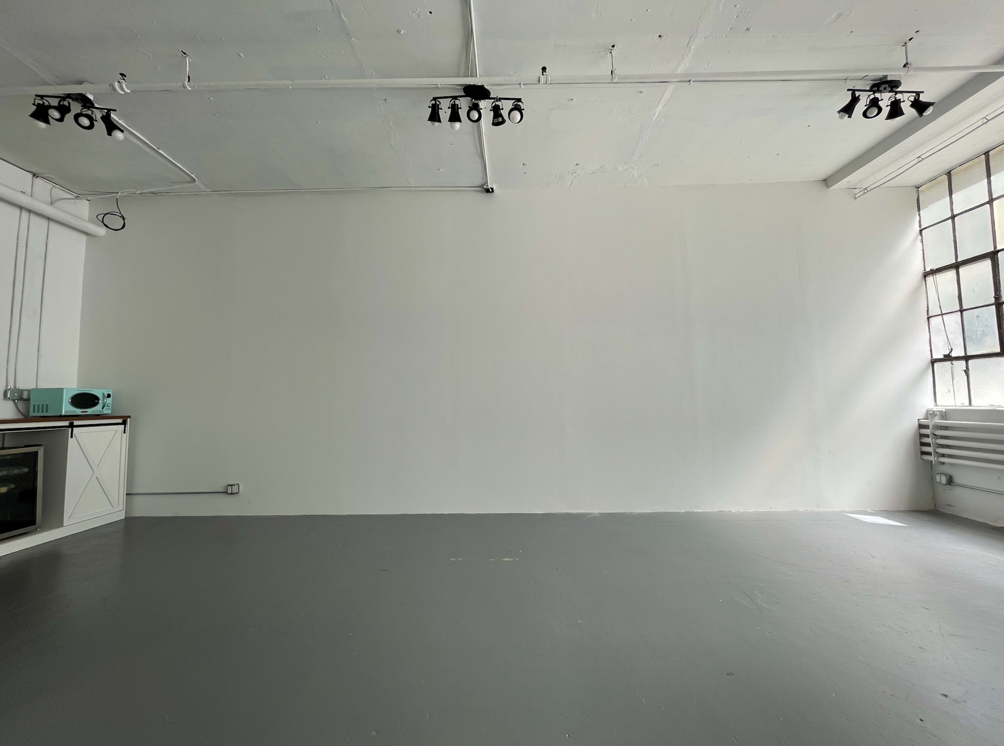 The image shows a vacant, well-lit room with white walls, large windows, and a gray floor, featuring minimal furnishings including a small teal appliance.