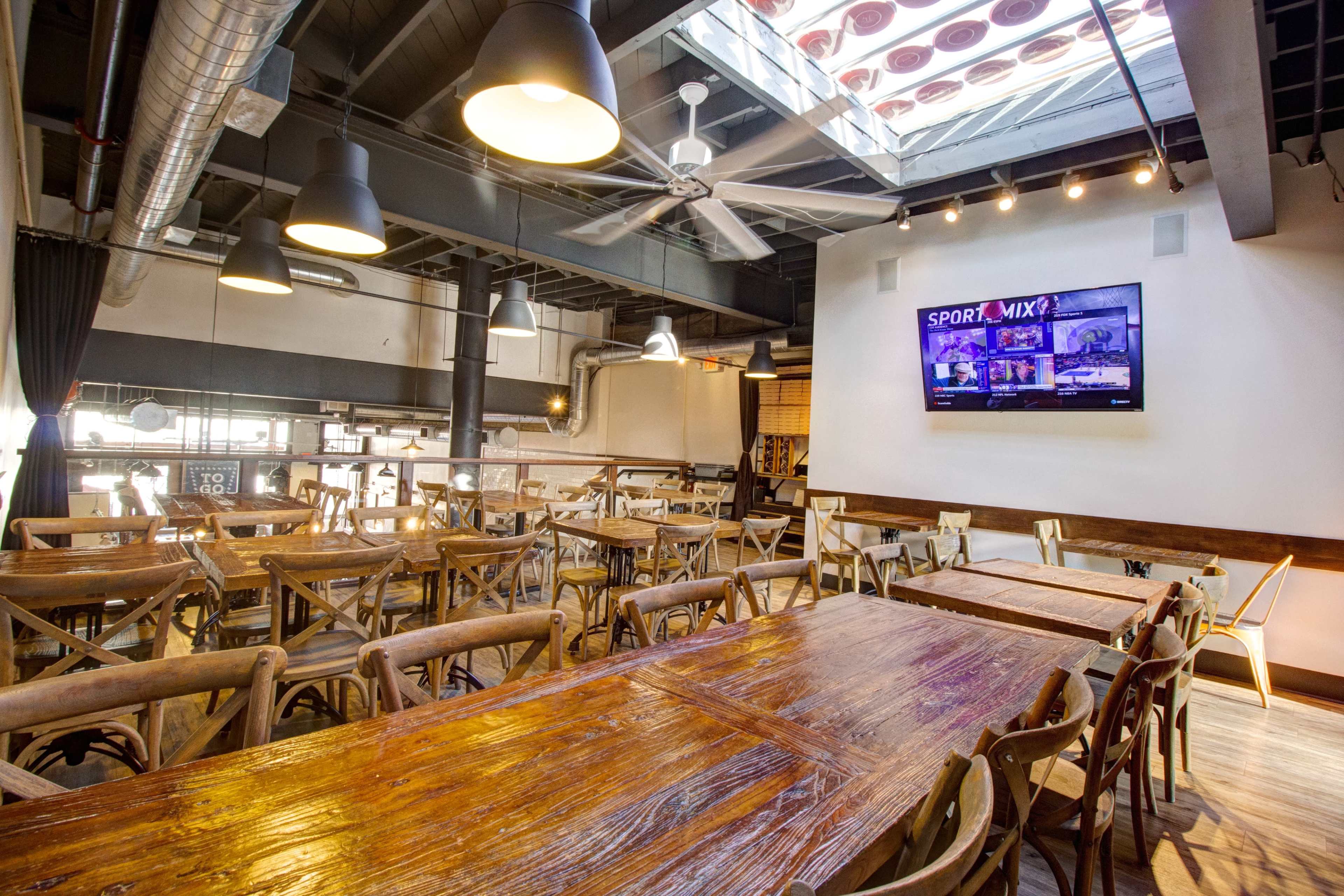 A modern restaurant interior with wooden tables arranged in rows, a large television mounted on the wall, and a skylight allowing natural light to enter.