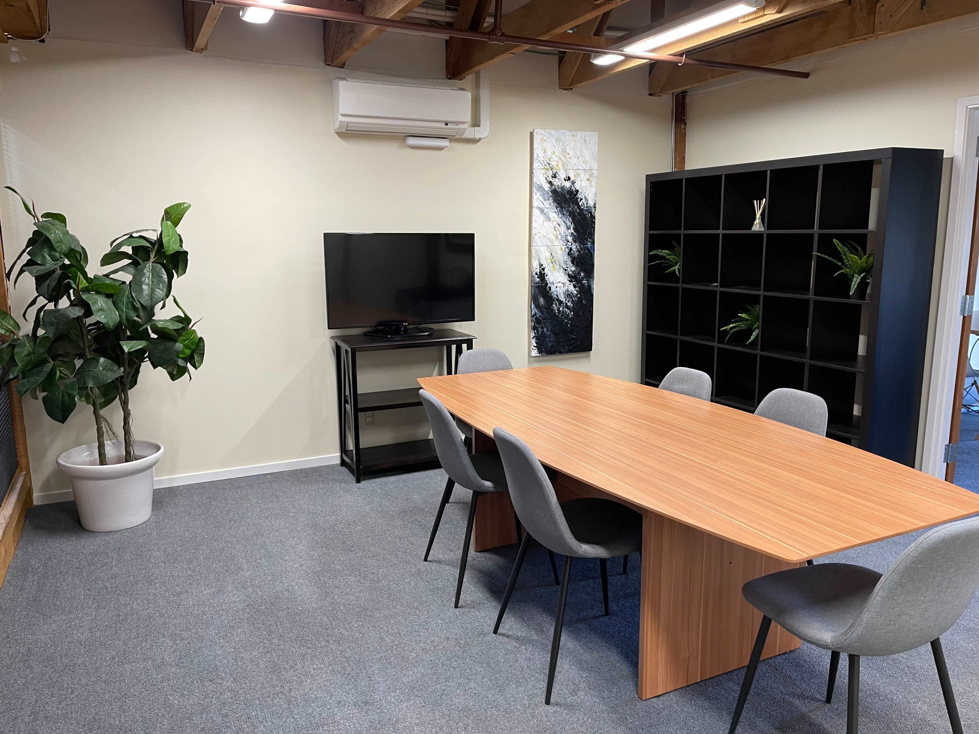 A modern meeting room featuring a wooden table surrounded by six chairs, with a television mounted on the wall and a tall plant in a pot nearby.