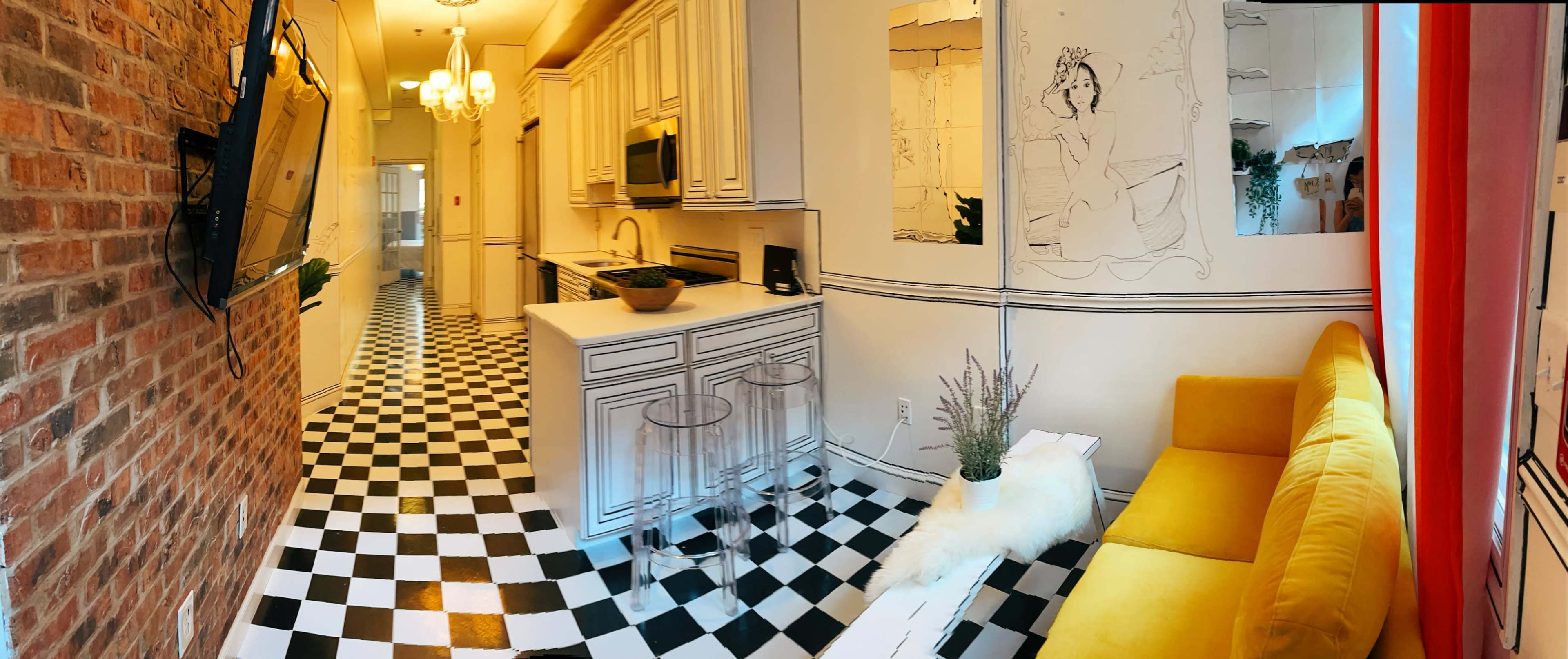 A modern living space with a yellow sofa, checkered black and white flooring, and a kitchen area featuring white cabinetry and a large television mounted on the wall.