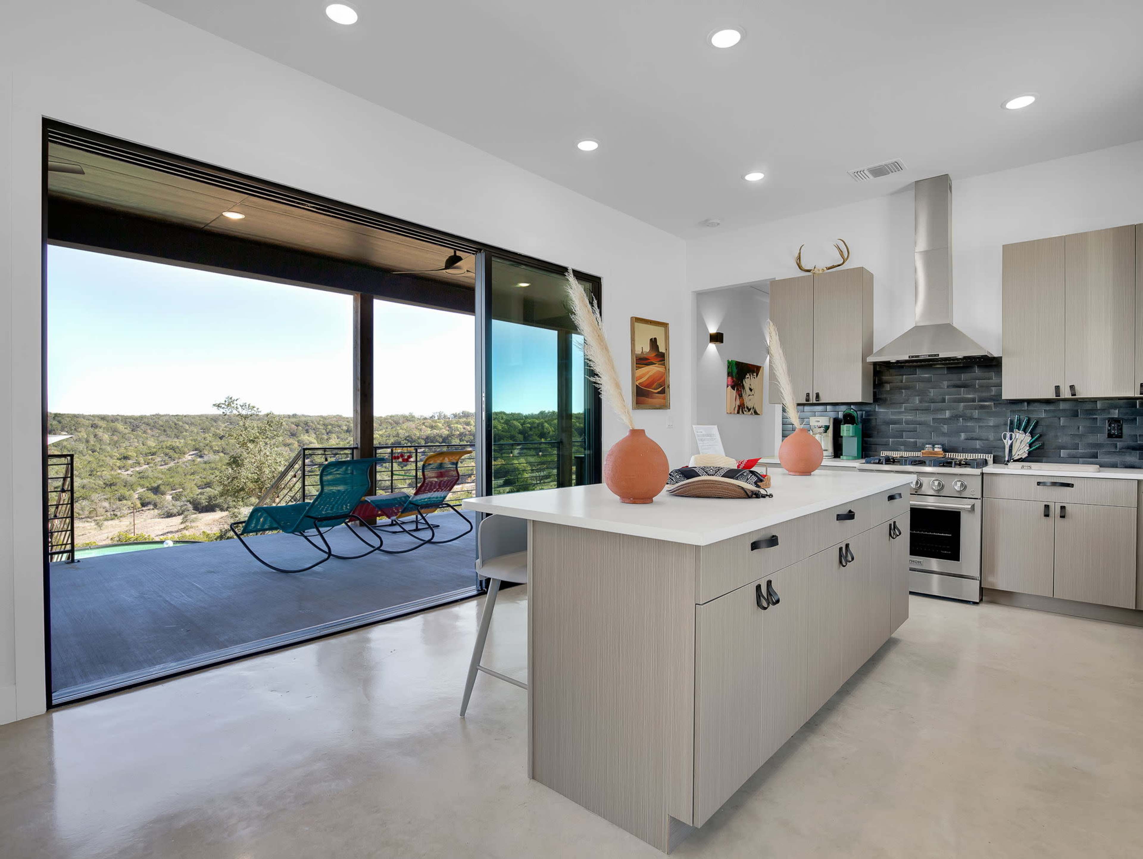 The image shows a modern kitchen with a central island, sleek cabinetry, and large windows opening to a scenic outdoor view.