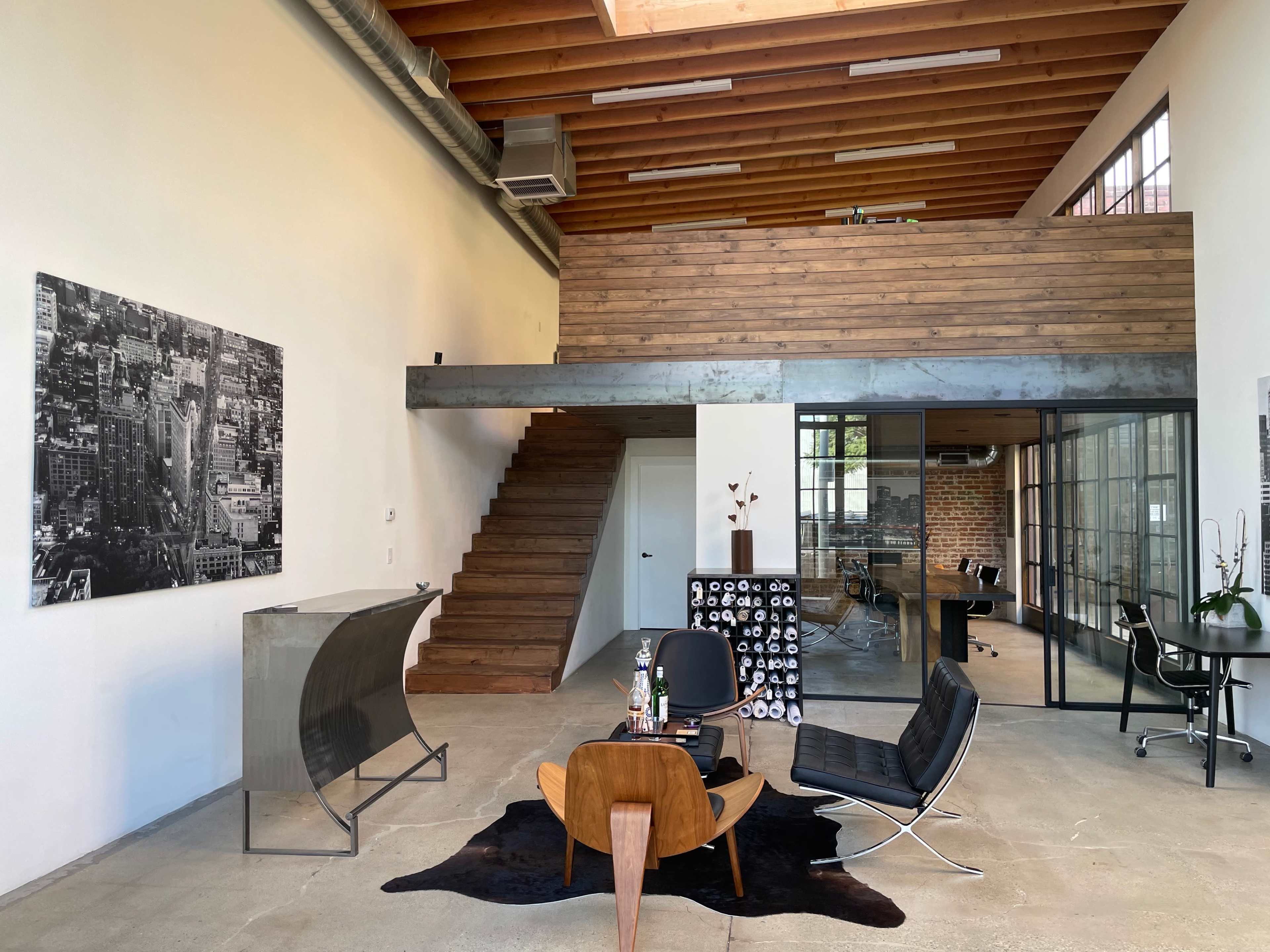 The image depicts a modern office space featuring a wooden staircase, an upstairs mezzanine area, and industrial-style furnishings.