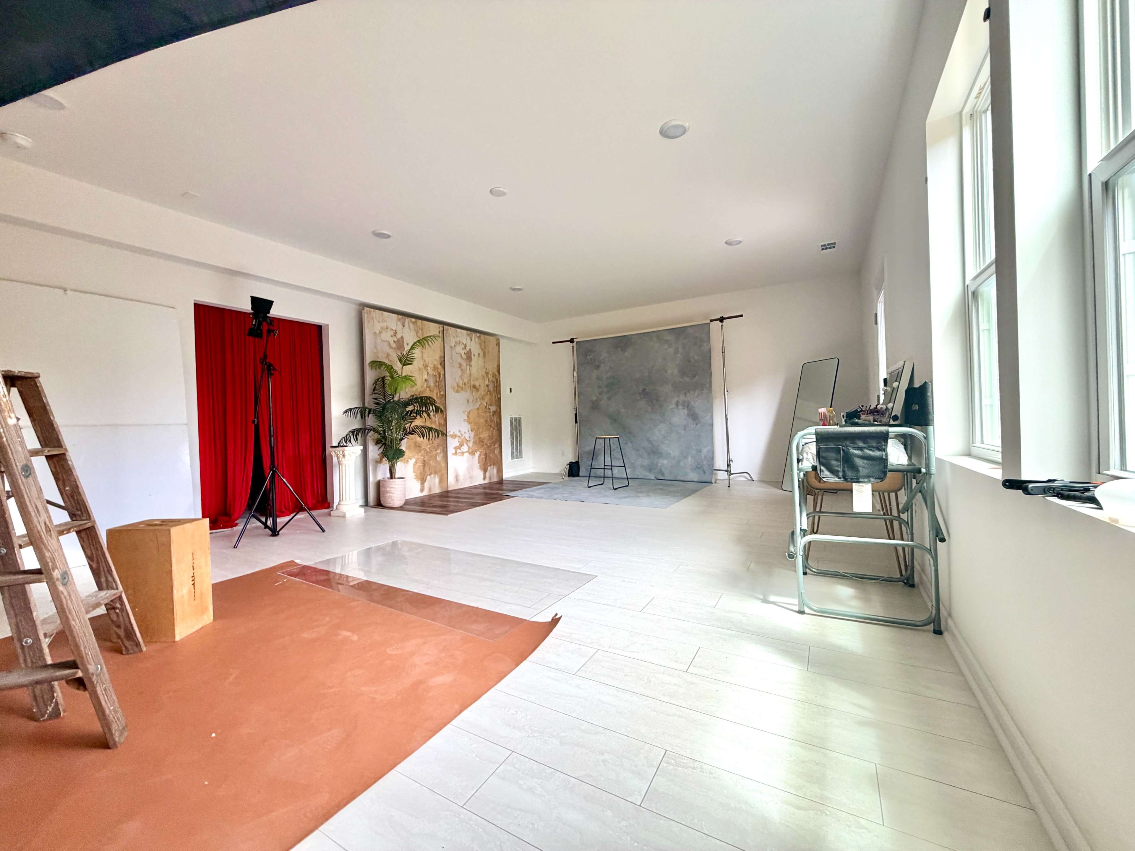 The image shows a spacious photography studio featuring a gray backdrop, red curtains, a ladder, and various props arranged around the room.