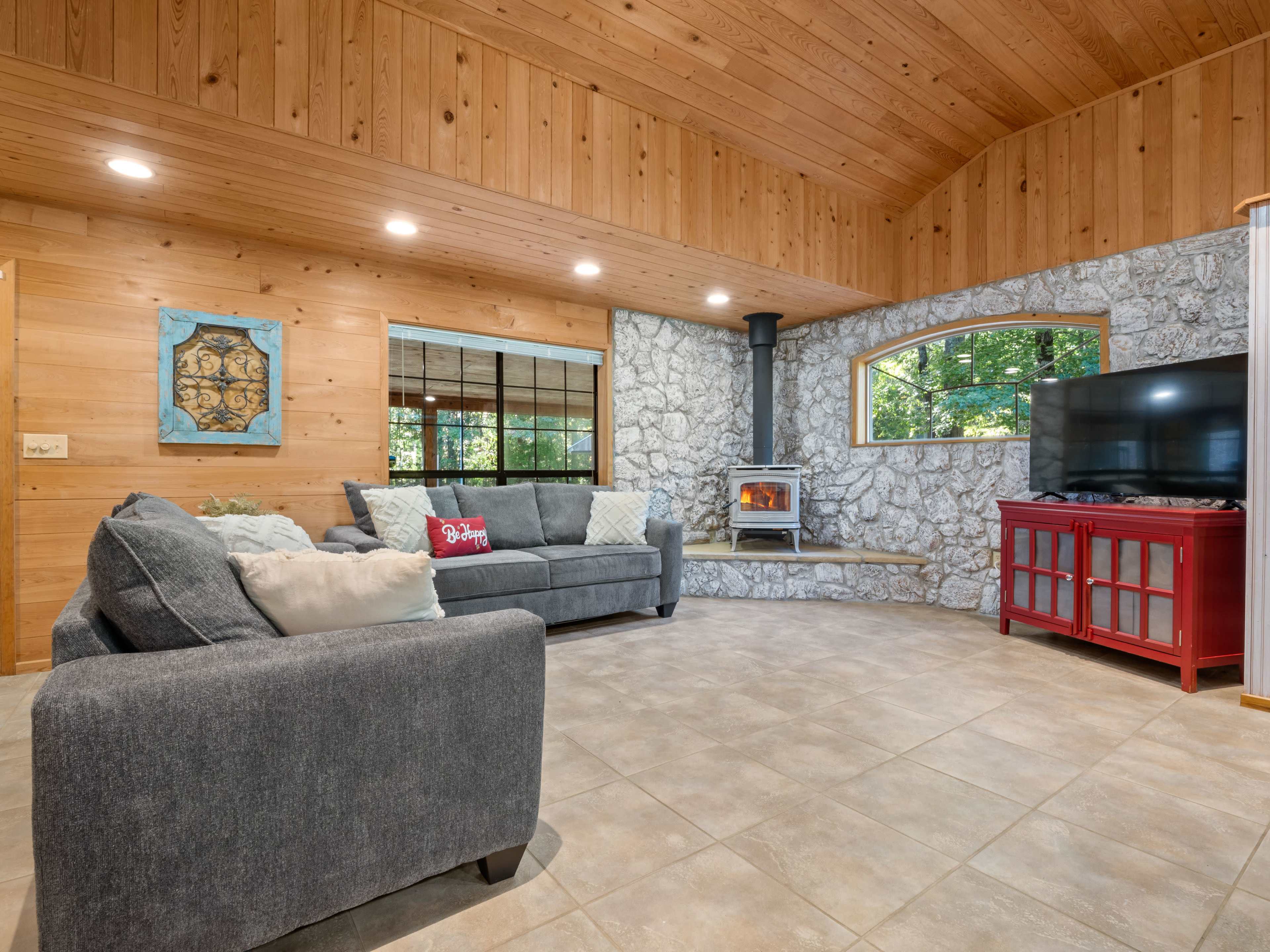 The cozy living room features a stone fireplace, a large sectional sofa, and a red low table, set against wooden walls and large windows letting in natural light.