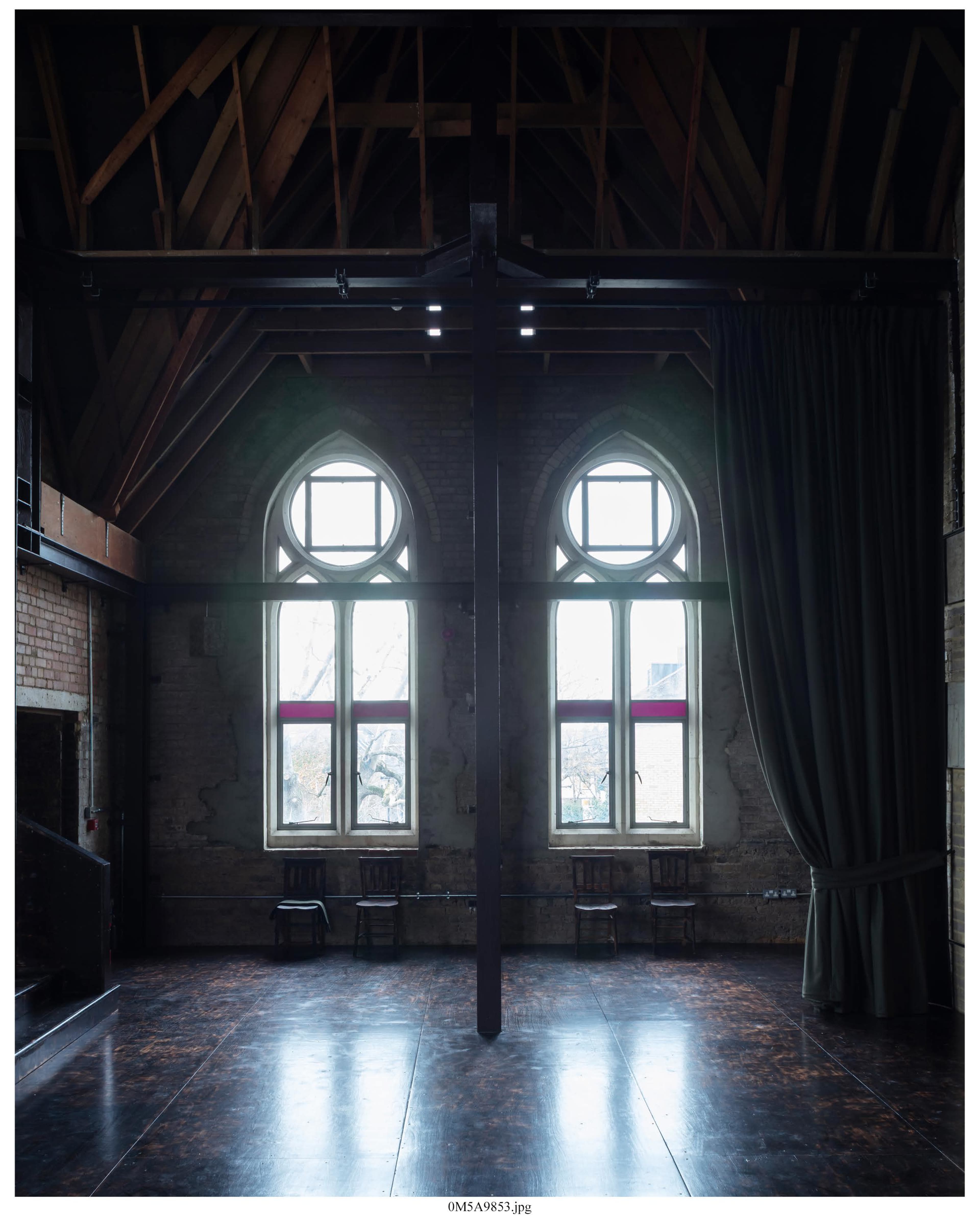 The image shows an empty interior space with wooden beams, large arched windows, and a dark curtain on one side.