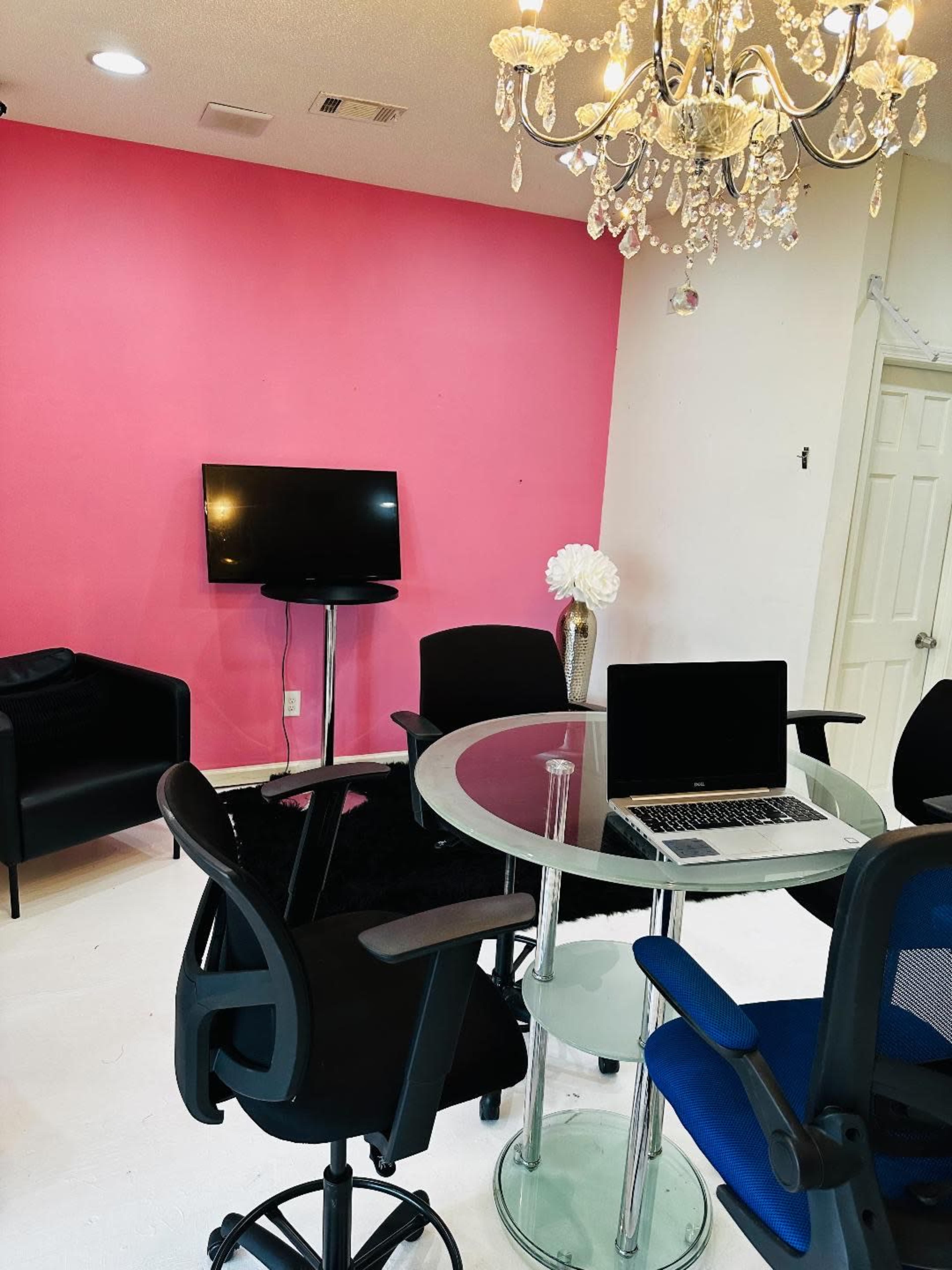A modern office space with a glass table, several chairs, a television mounted on a pink wall, and a chandelier overhead.