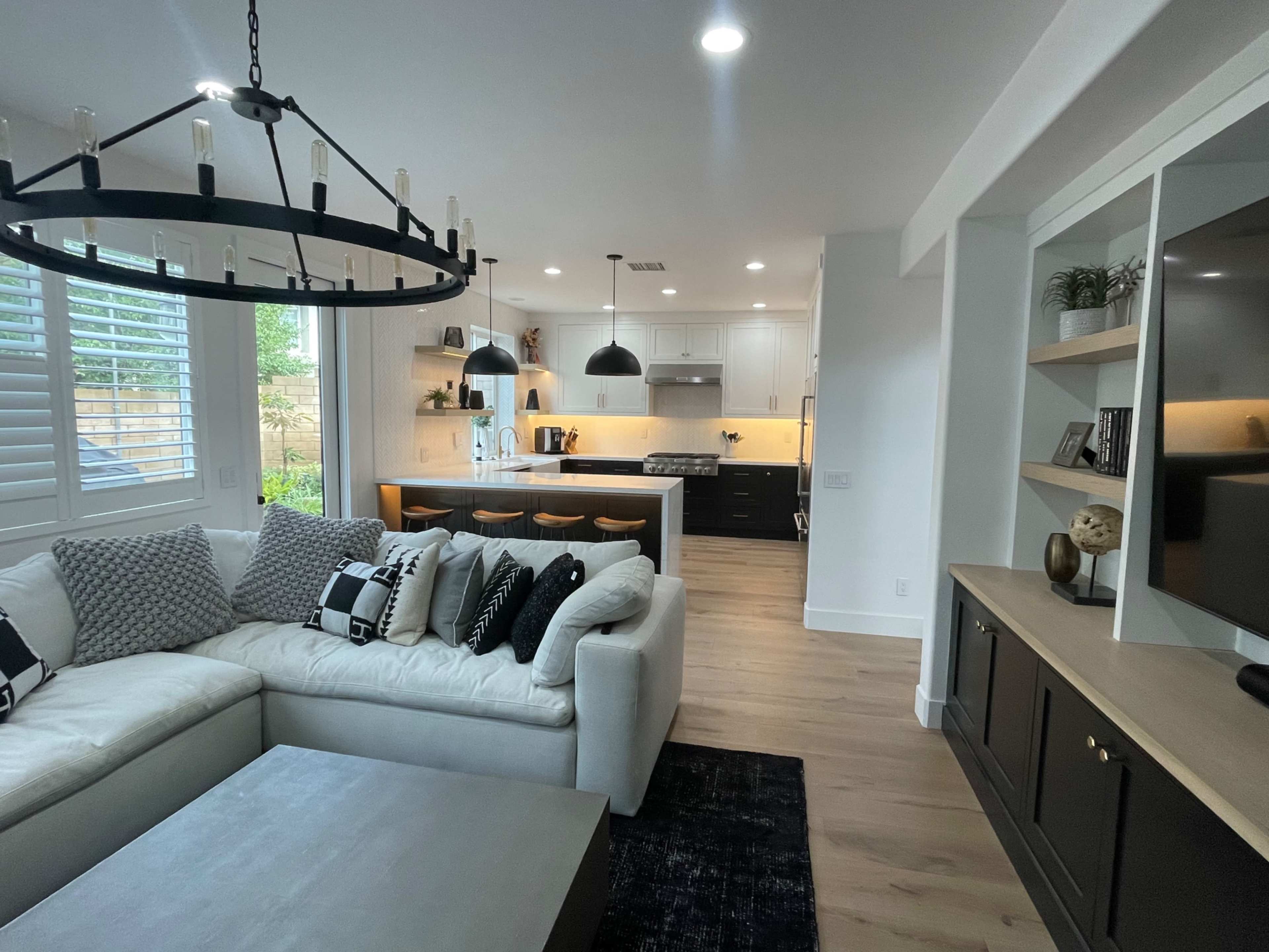 A modern living room with a large white sectional sofa, a coffee table, and a kitchen in the background featuring dark cabinets and a central island with bar stools.