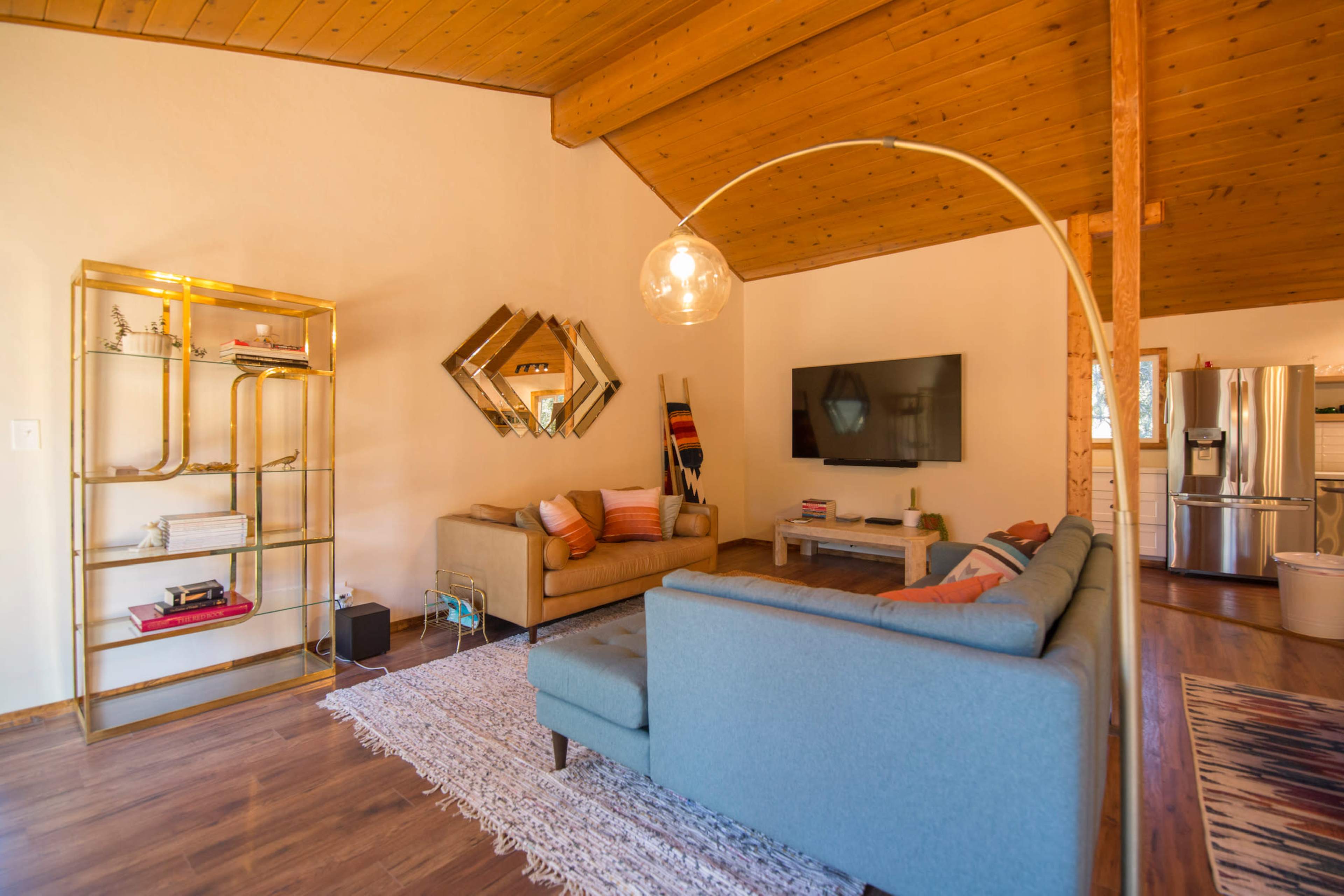 The living room features a beige sofa, a light blue sectional, a gold shelving unit, and a large wall-mounted television, all under a wooden ceiling.