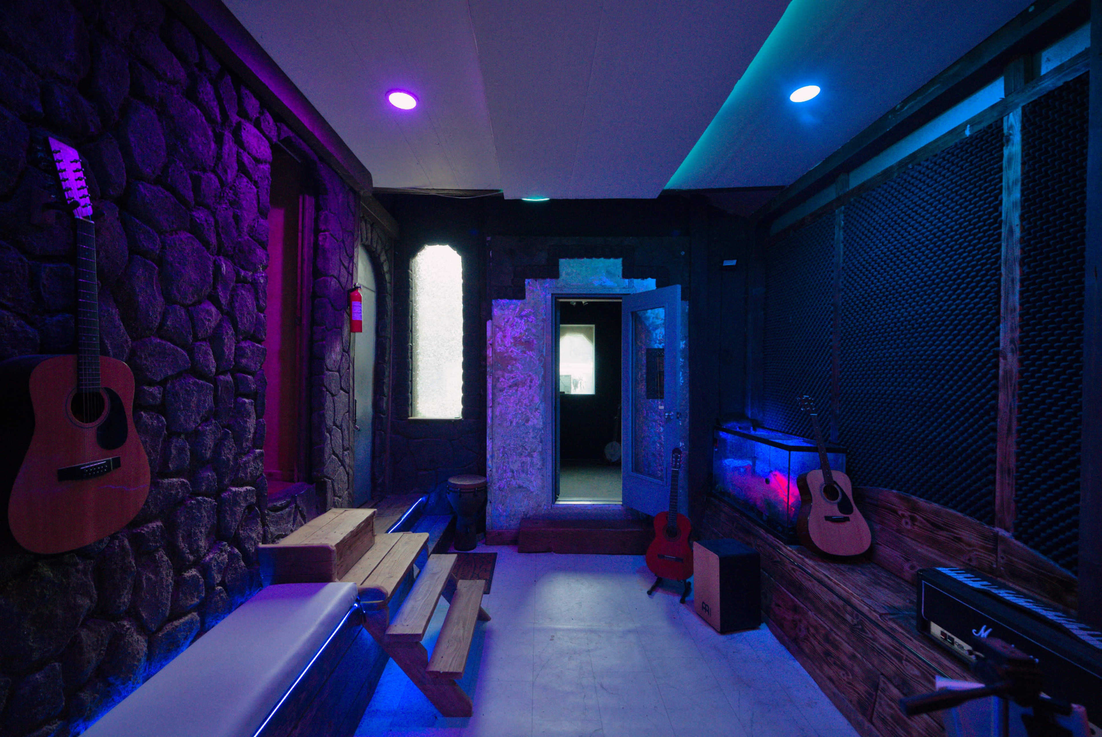 The image depicts a dimly lit room with stone walls, featuring wooden benches, several guitars on display, and a door leading to a brightly lit hallway.