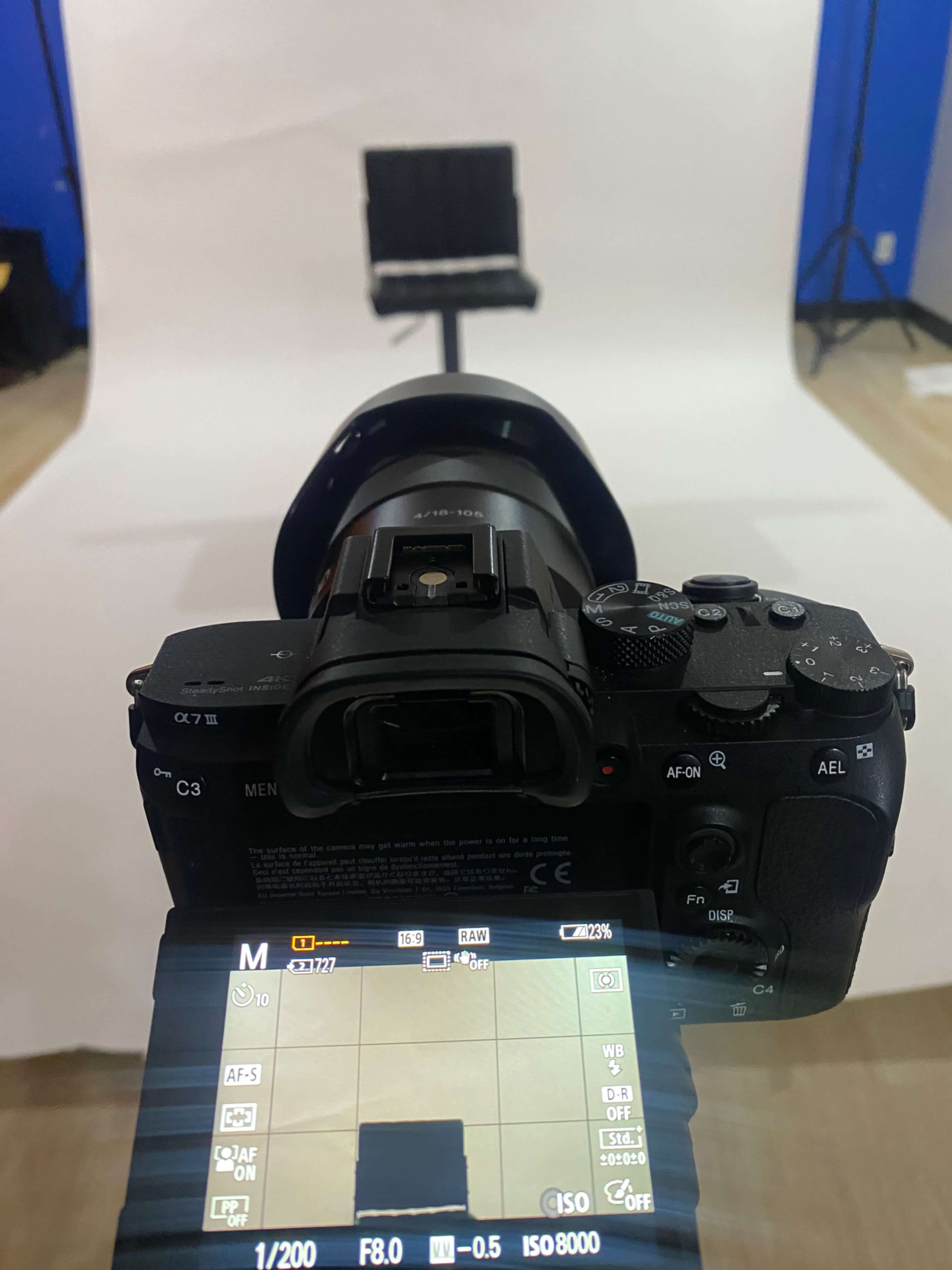 A camera is positioned on a tripod, facing a black chair against a white backdrop in a photography studio.