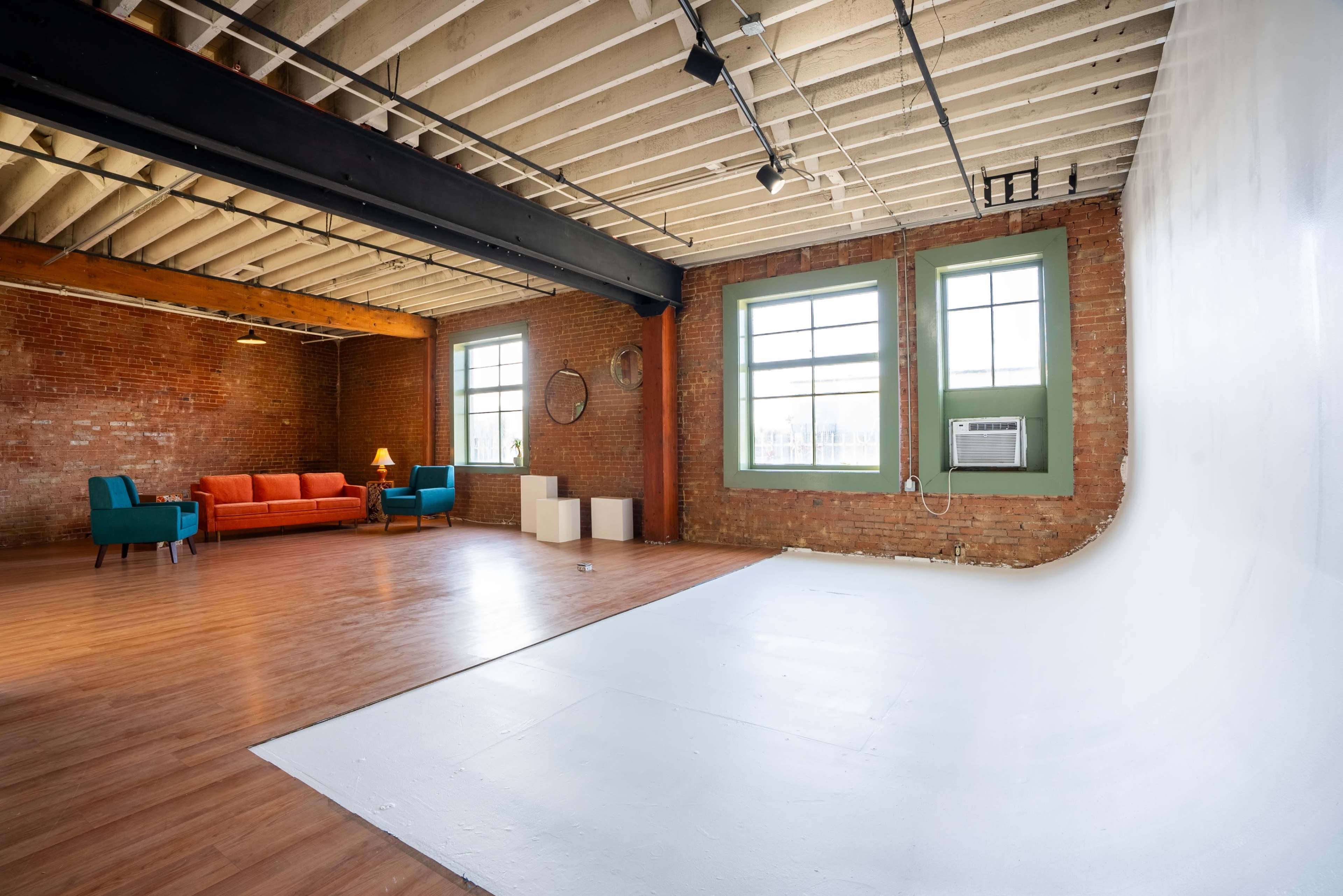 The image shows a spacious, industrial-style room featuring exposed brick walls, wooden flooring, and large windows, with a brightly lit area set up for photography.