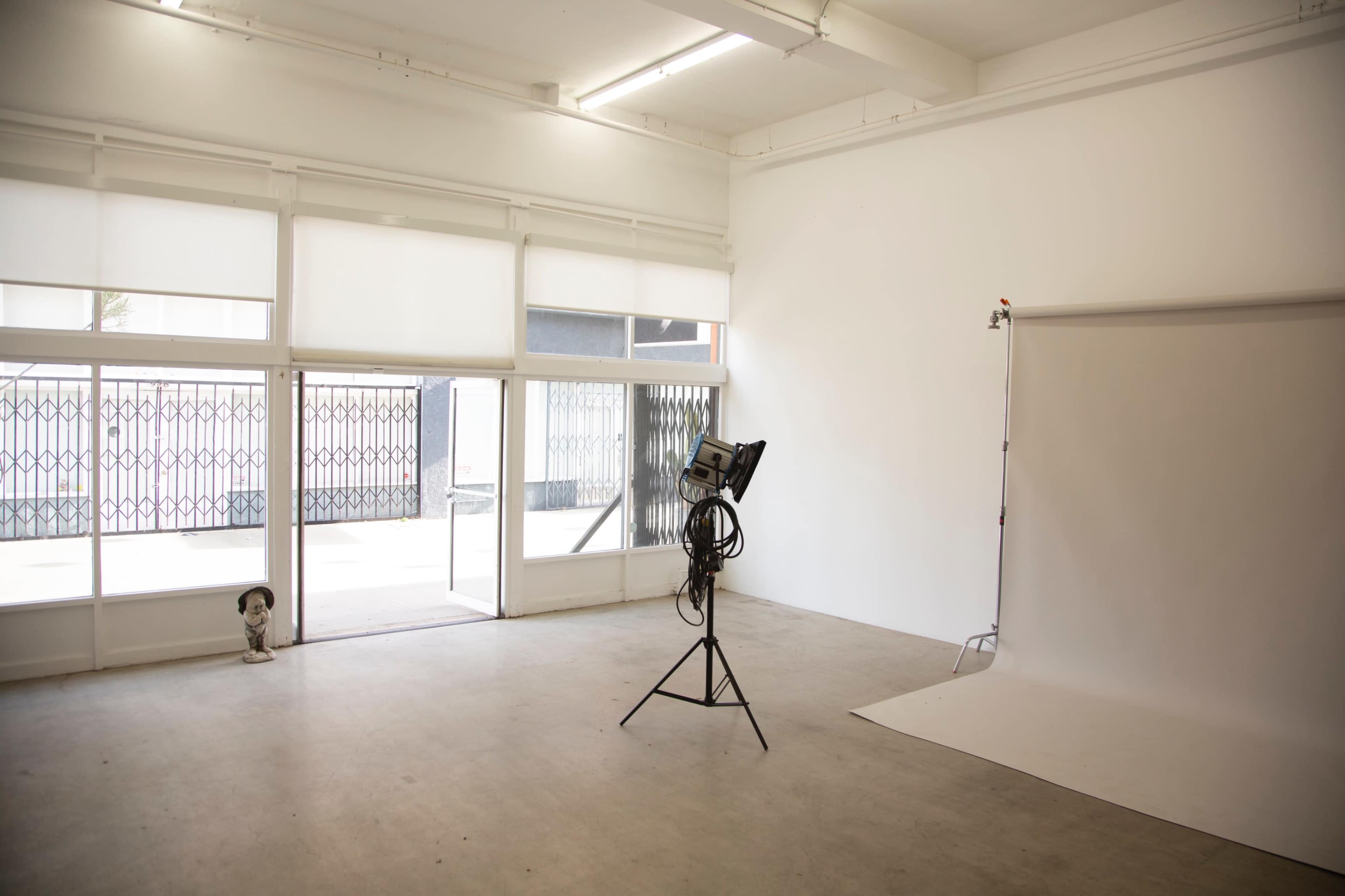 A spacious, empty studio with large windows, a light on a stand, and a white backdrop against the wall.