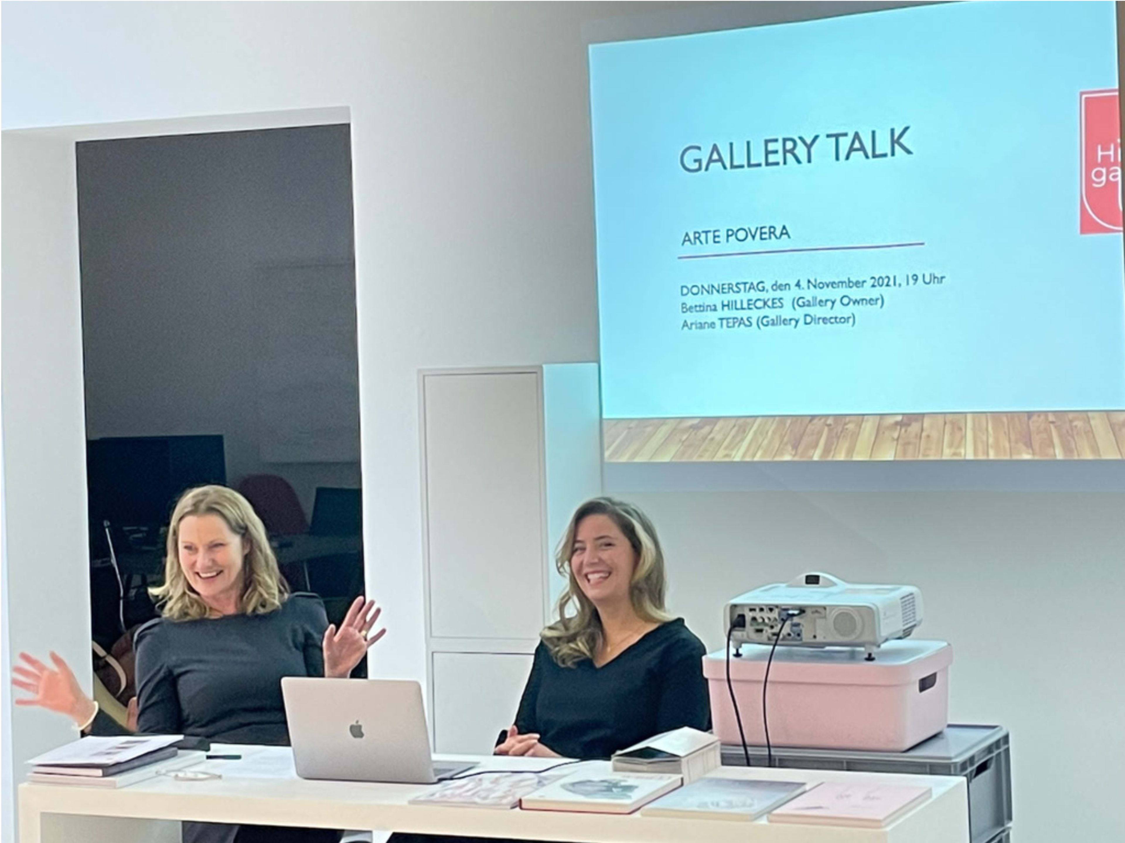 Two women are seated at a table during a gallery talk event with a presentation slide about "Arte Povera" displayed behind them.