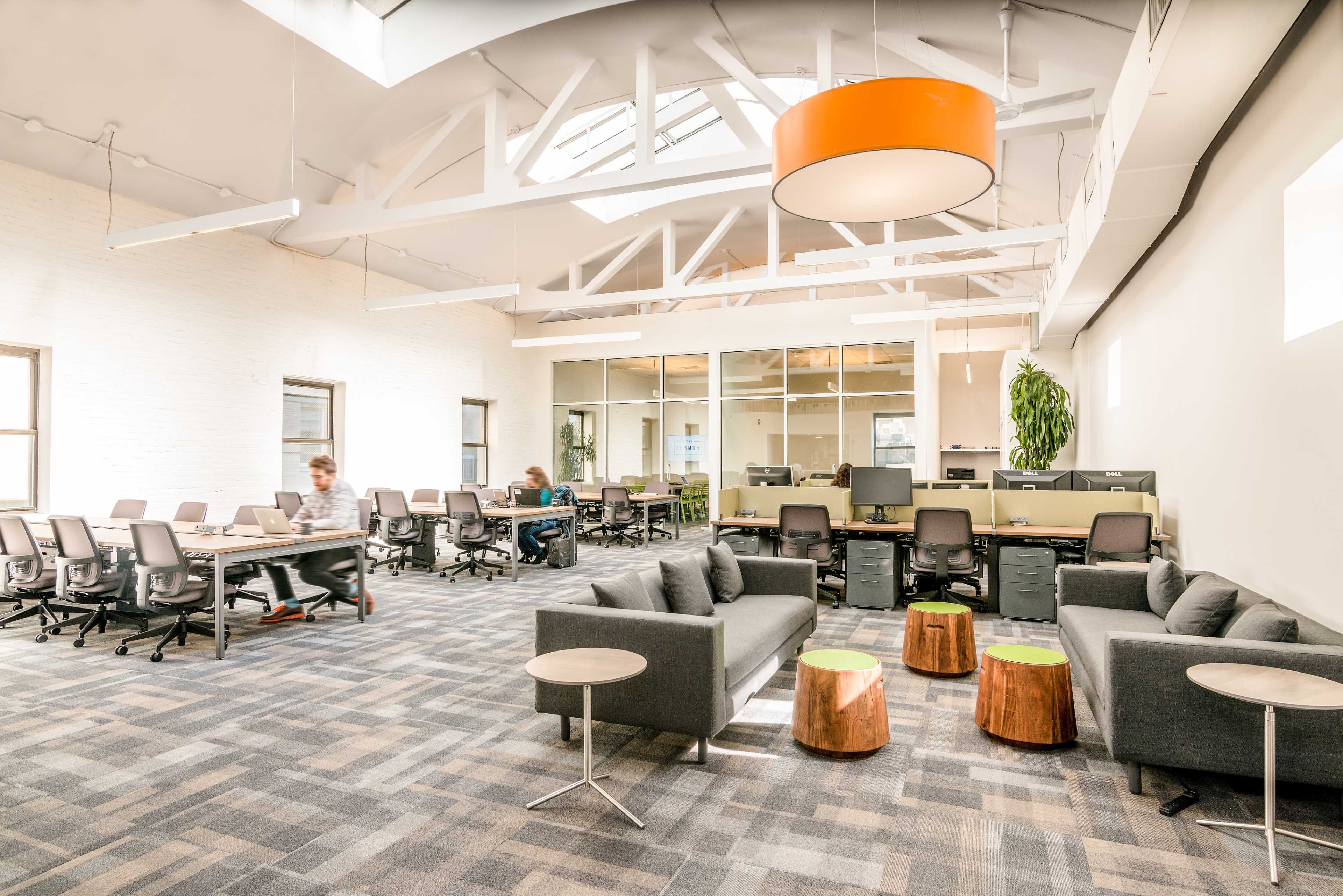 A modern office space featuring a lounge area with gray sofas and wooden stools, alongside desks equipped with computers and large windows letting in natural light.