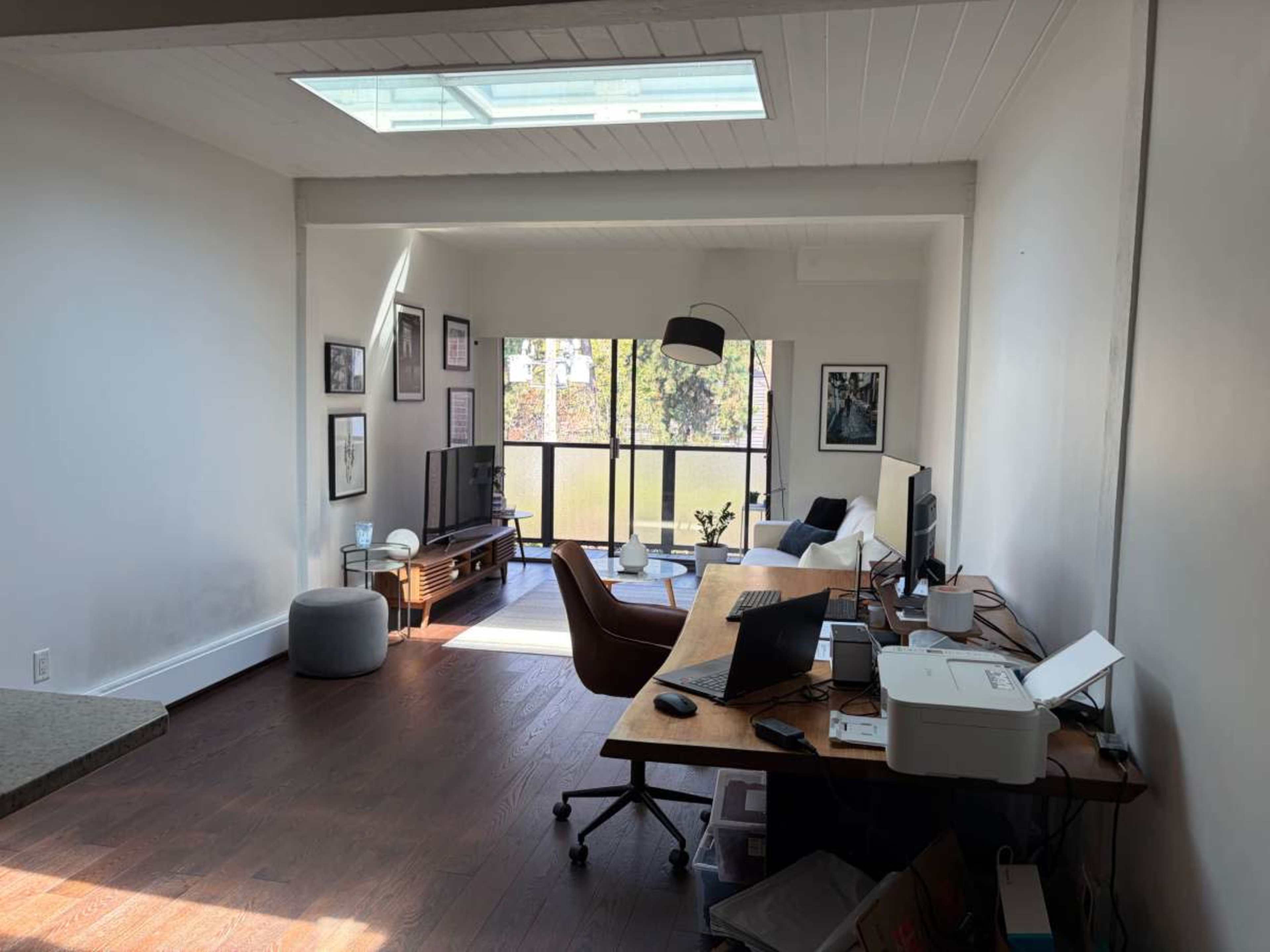The image shows a modern, open living space featuring a workspace with a desk and computer, a seating area, and large windows overlooking a view of trees.