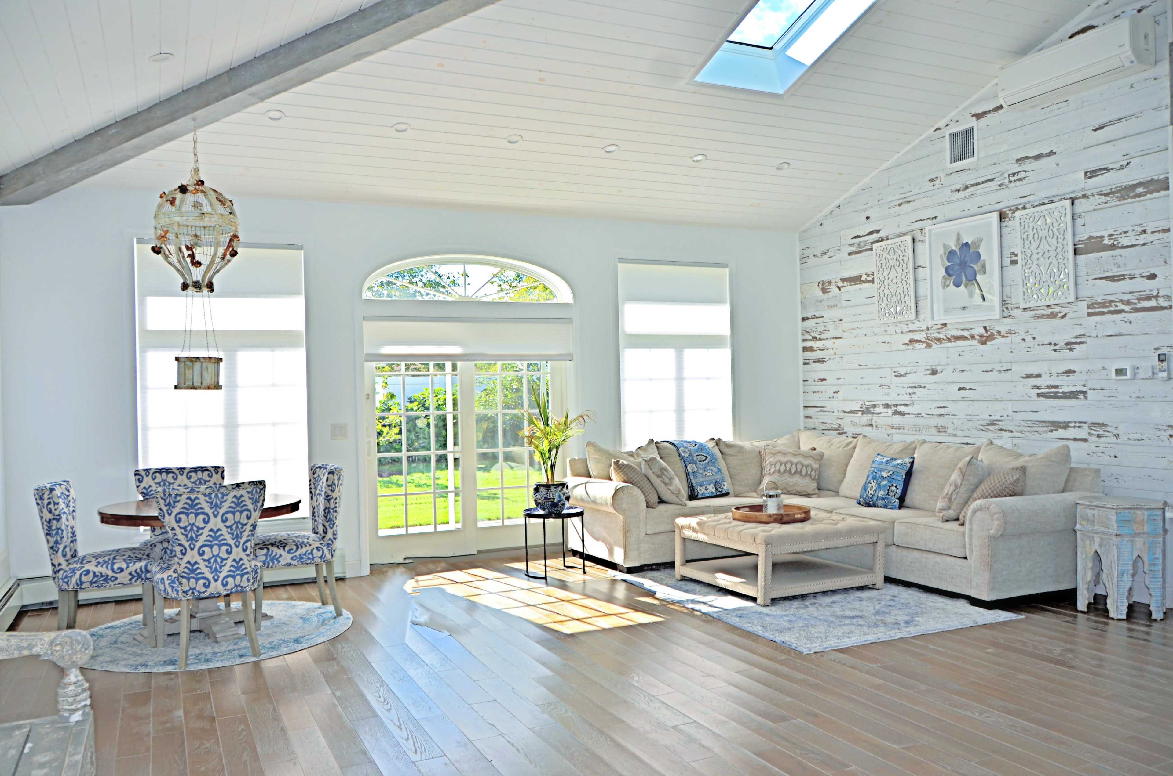 A bright living room features a comfortable sofa, a wooden coffee table, and a dining set, illuminated by natural light streaming through large windows.