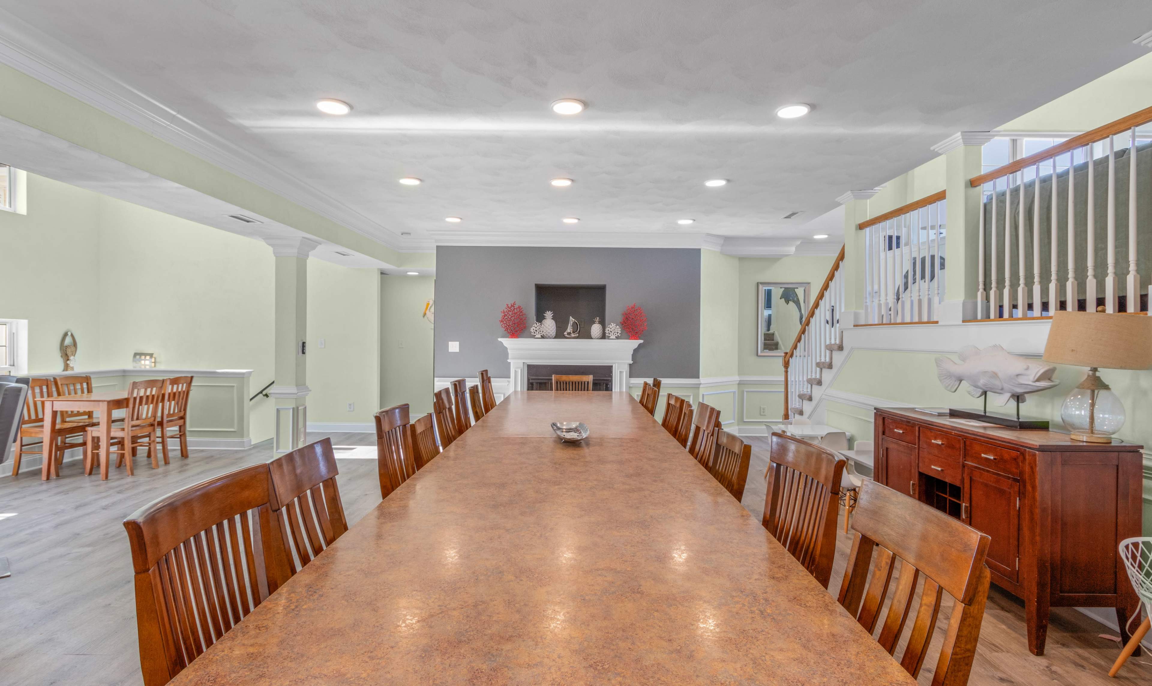 A spacious dining area featuring a long table with wooden chairs, a fireplace against a gray wall, and a staircase leading to an upper level.