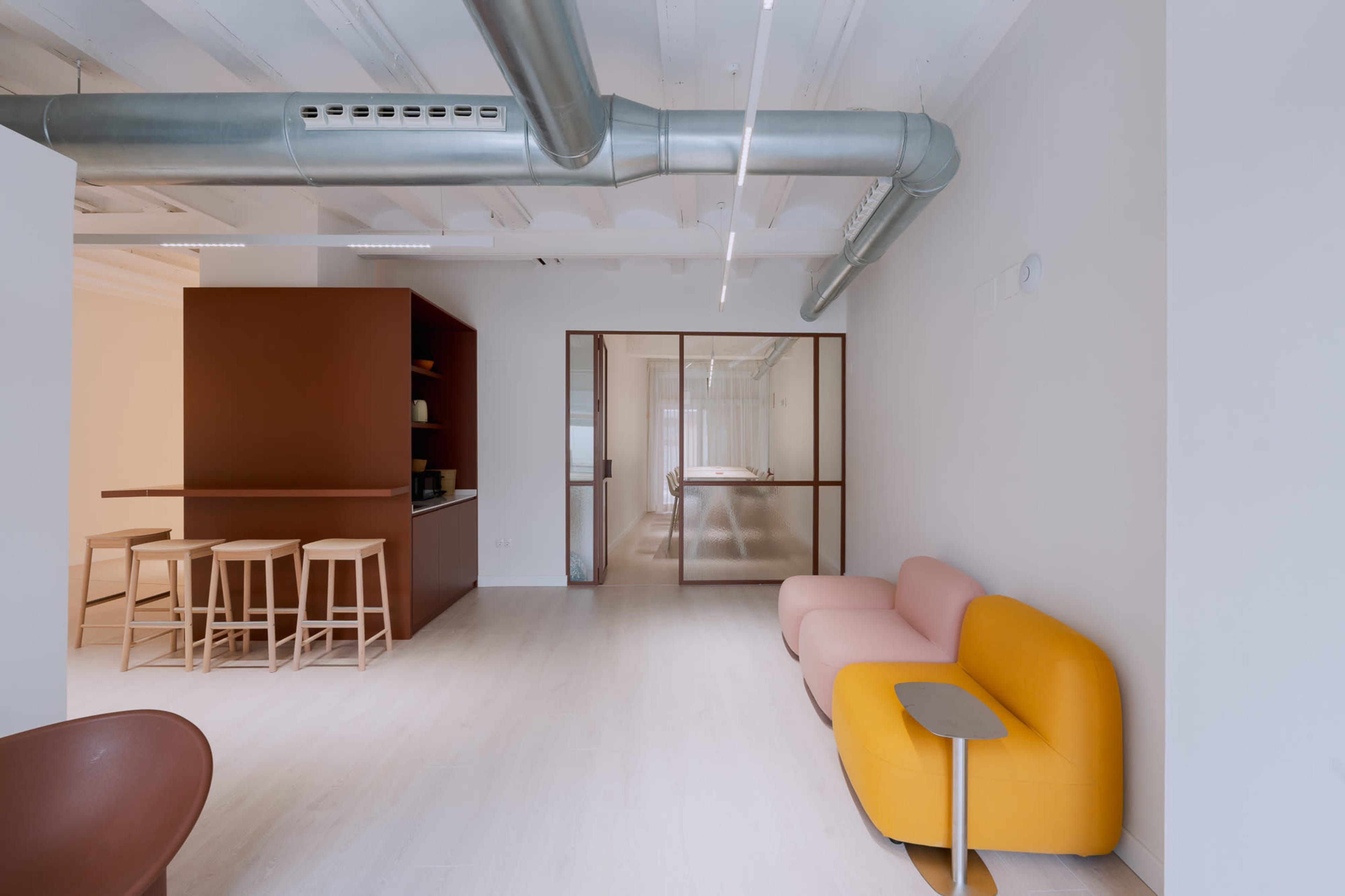 The image shows a modern, minimalist office space featuring a pink and yellow sofa, wooden bar stools, and exposed metal ductwork.