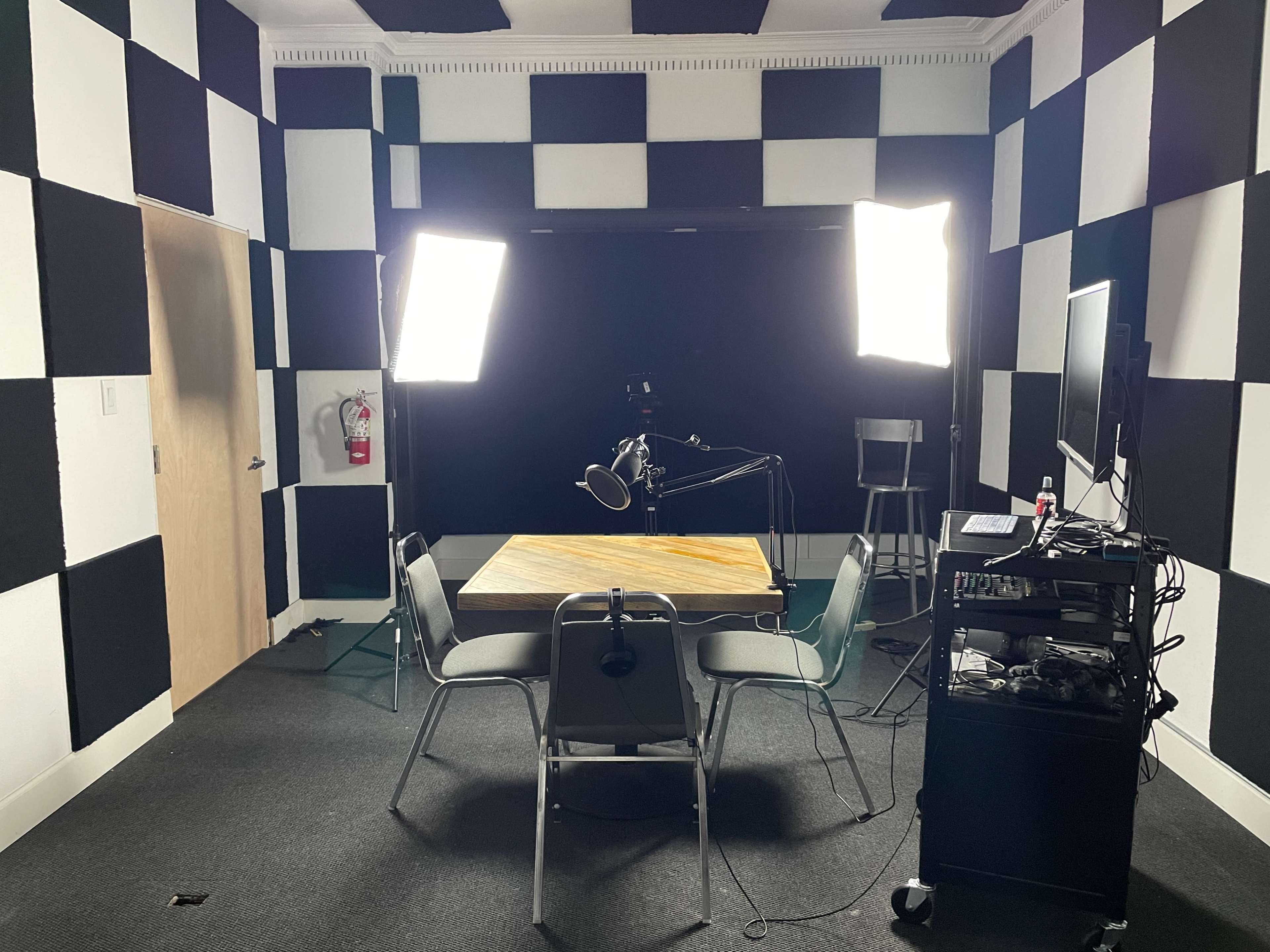 A recording studio features a central table surrounded by chairs, with two bright lights positioned to illuminate a black backdrop and various equipment on a nearby cart.