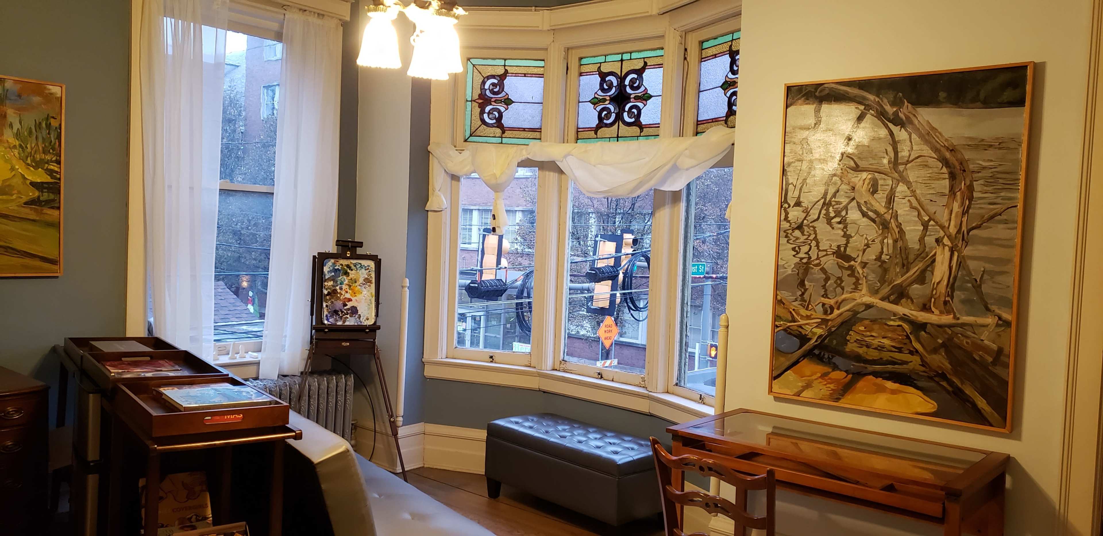 A room with large windows featuring stained glass, a small seating area, and various artworks displayed on the walls.
