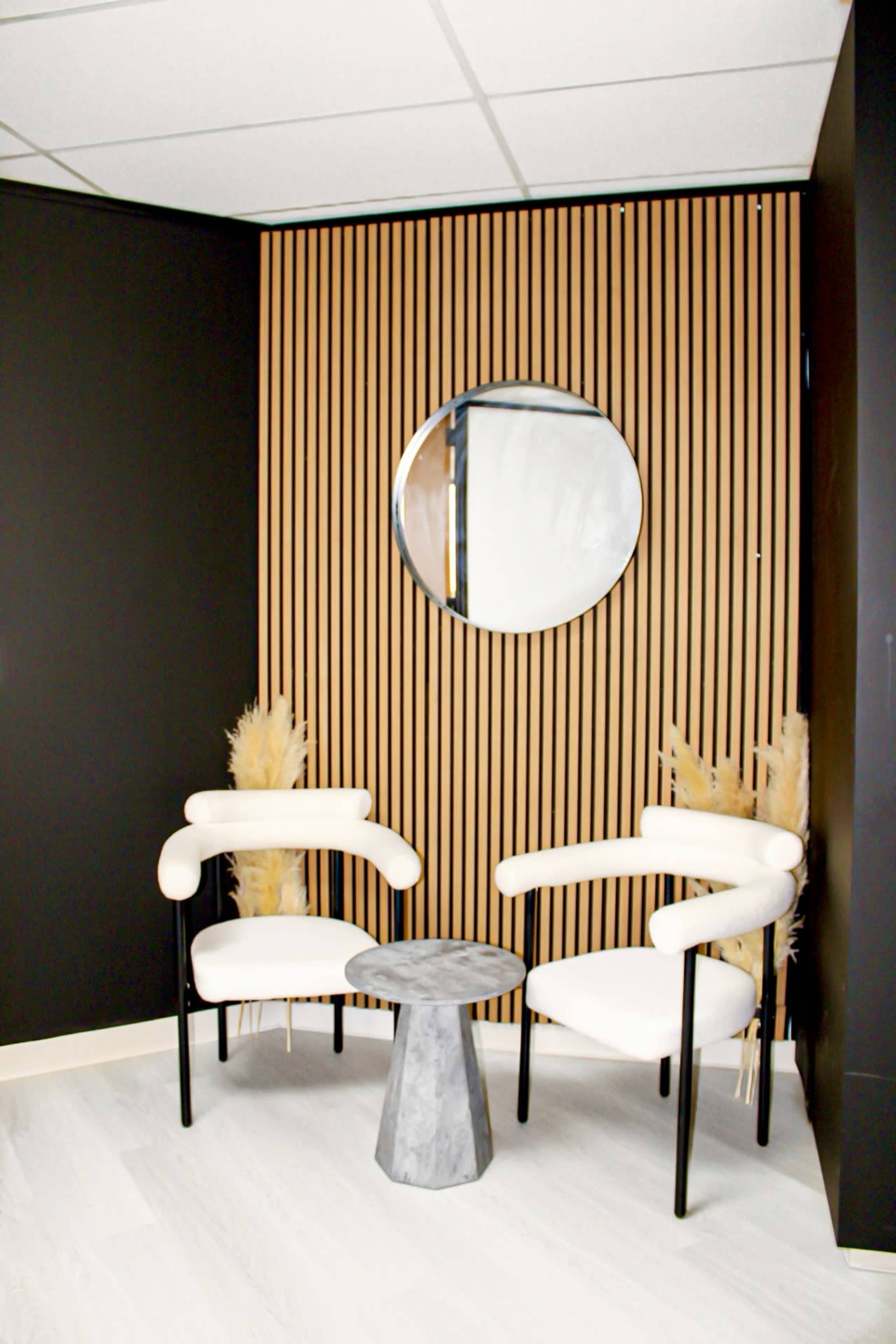 The image shows a cozy seating area featuring two modern white chairs and a small gray marble table against a backdrop of wooden slats and a round mirror.