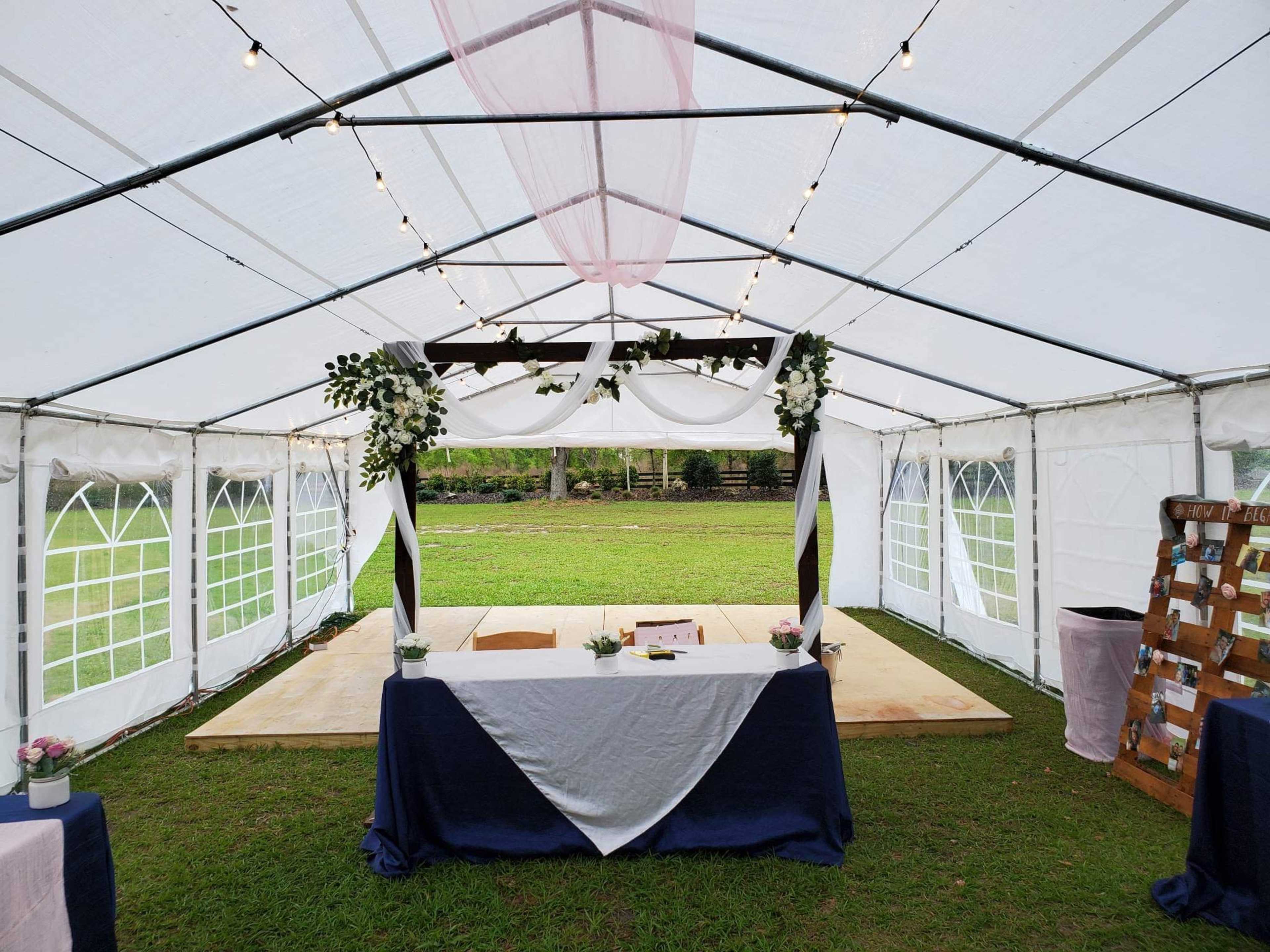 A tented wedding reception area features a decorated table at the front with floral arrangements and a backdrop of greenery.