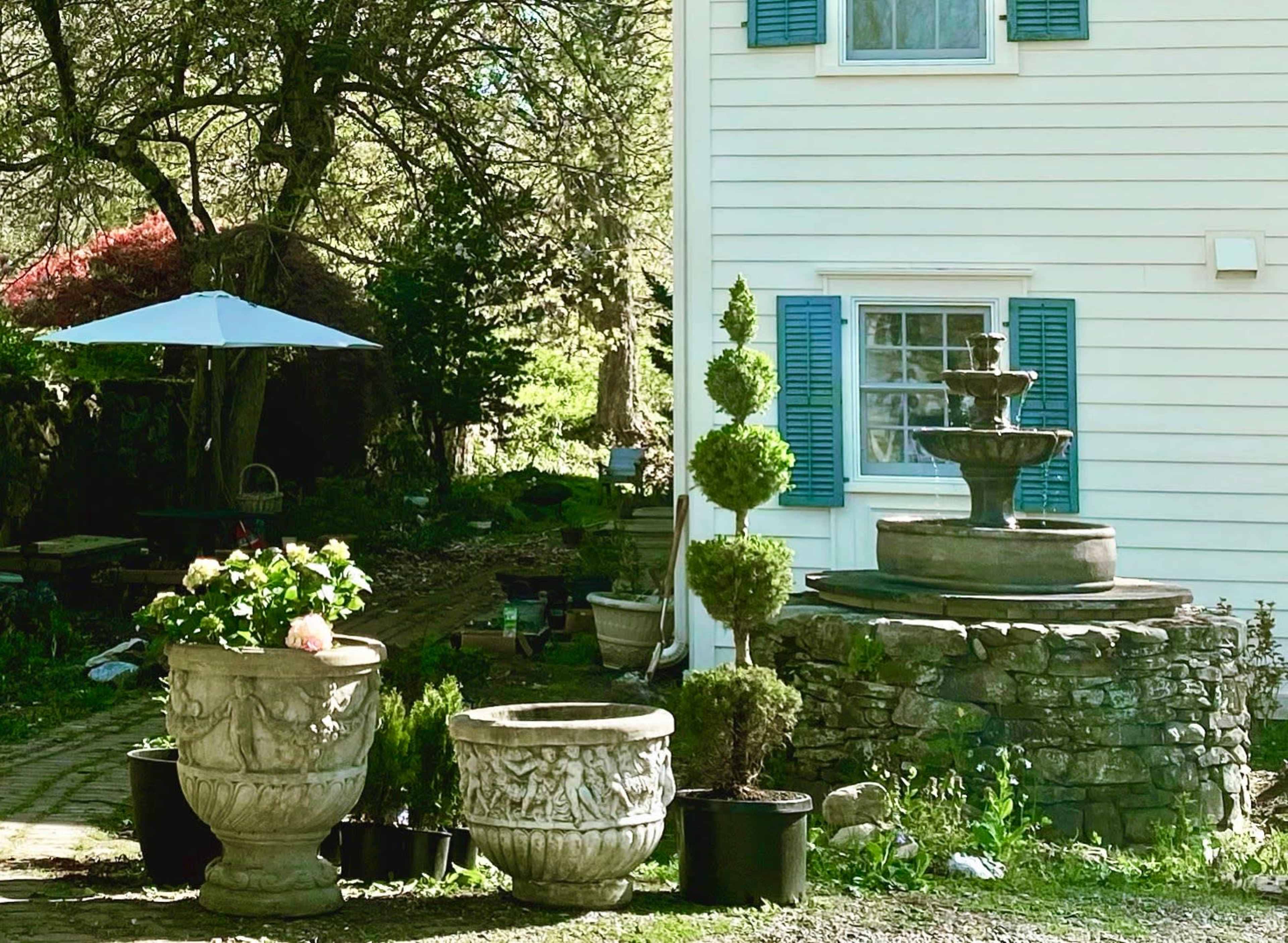 A tiered stone fountain stands next to two decorative planters in a garden area beside a white house.