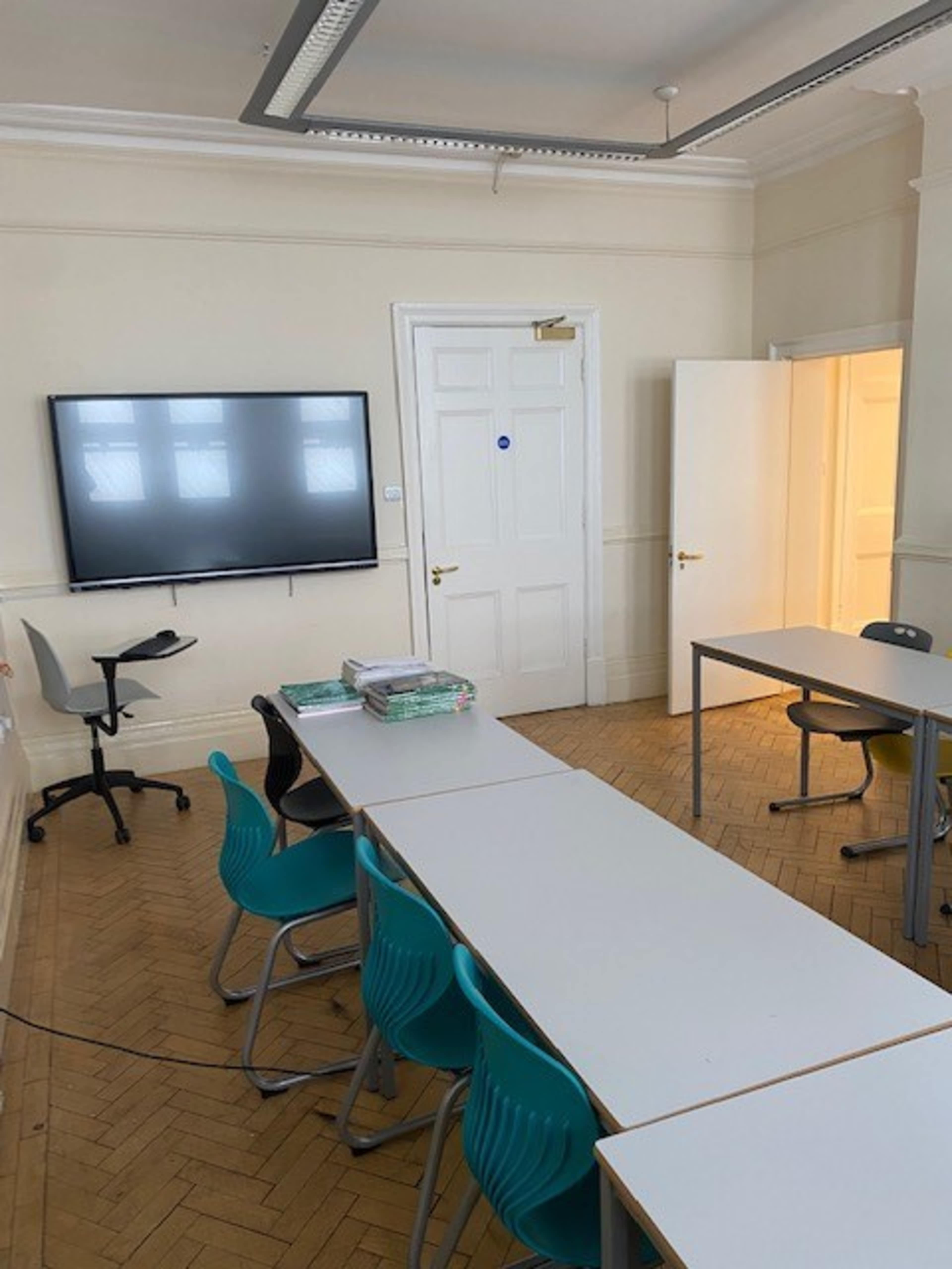 A classroom is set up with a long table, several blue chairs, a wall-mounted screen, and an open door leading to another room.