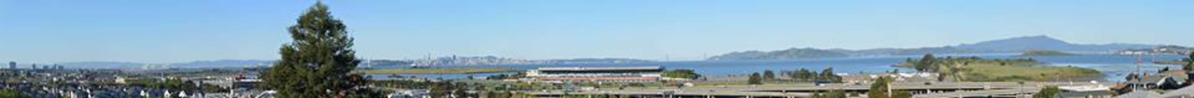 The image shows a panoramic view of a waterfront area with a large body of water, distant hills, and urban buildings.