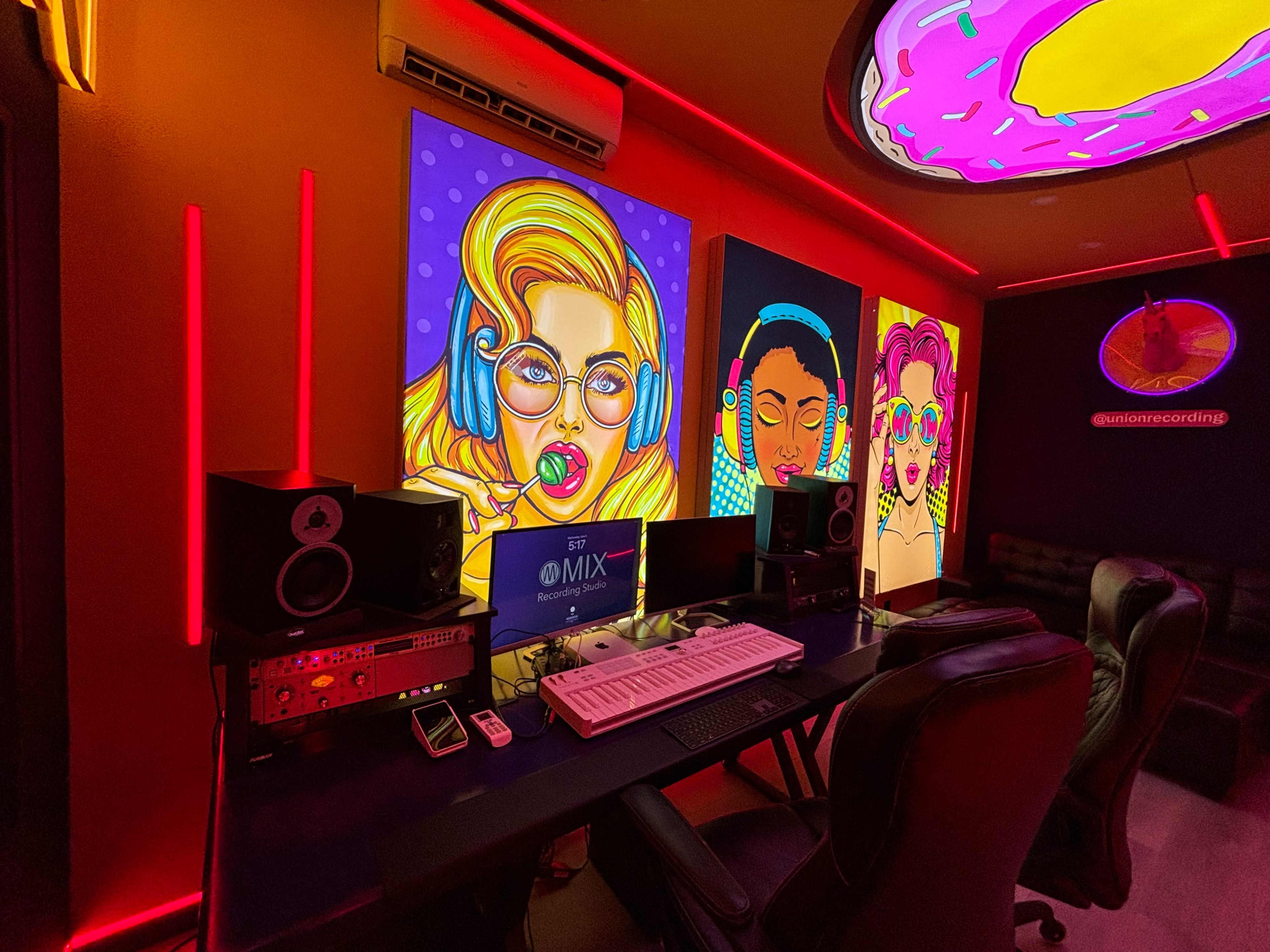 A music recording studio featuring colorful pop art portraits of women on the walls, with a mixing console, speakers, and a computer setup on a desk.