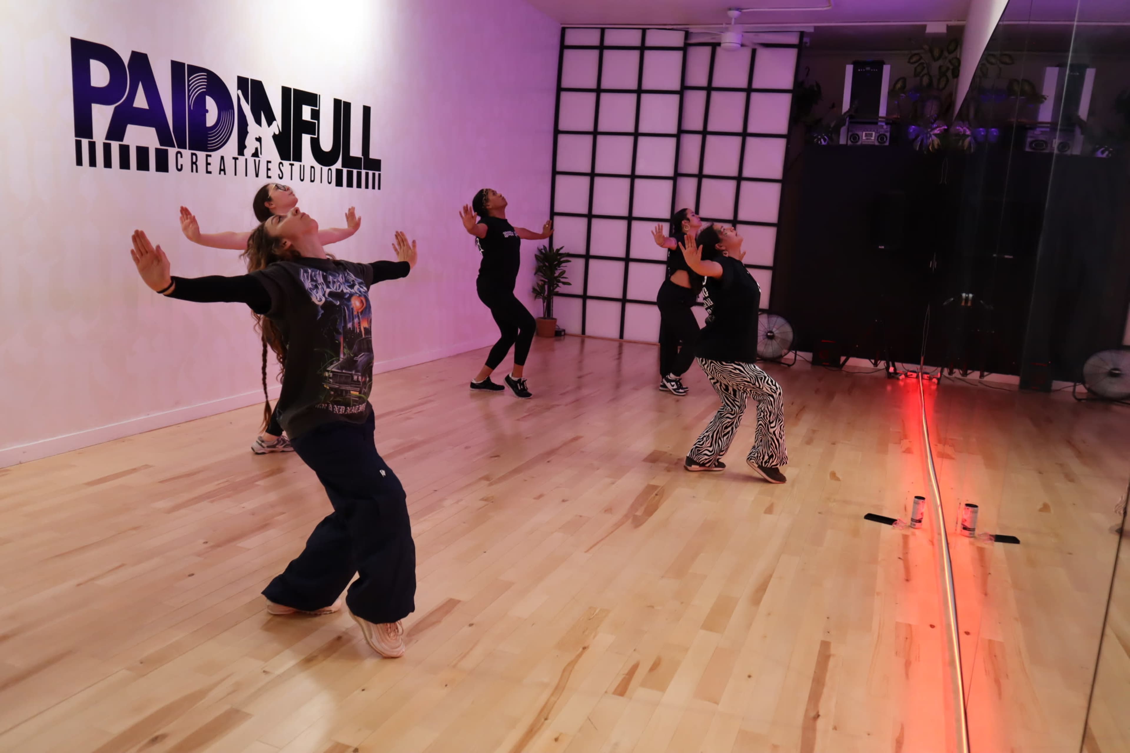A group of five people is dancing in a studio with mirrored walls and wooden flooring, while the words "PAID IN FULL" are displayed on the back wall.