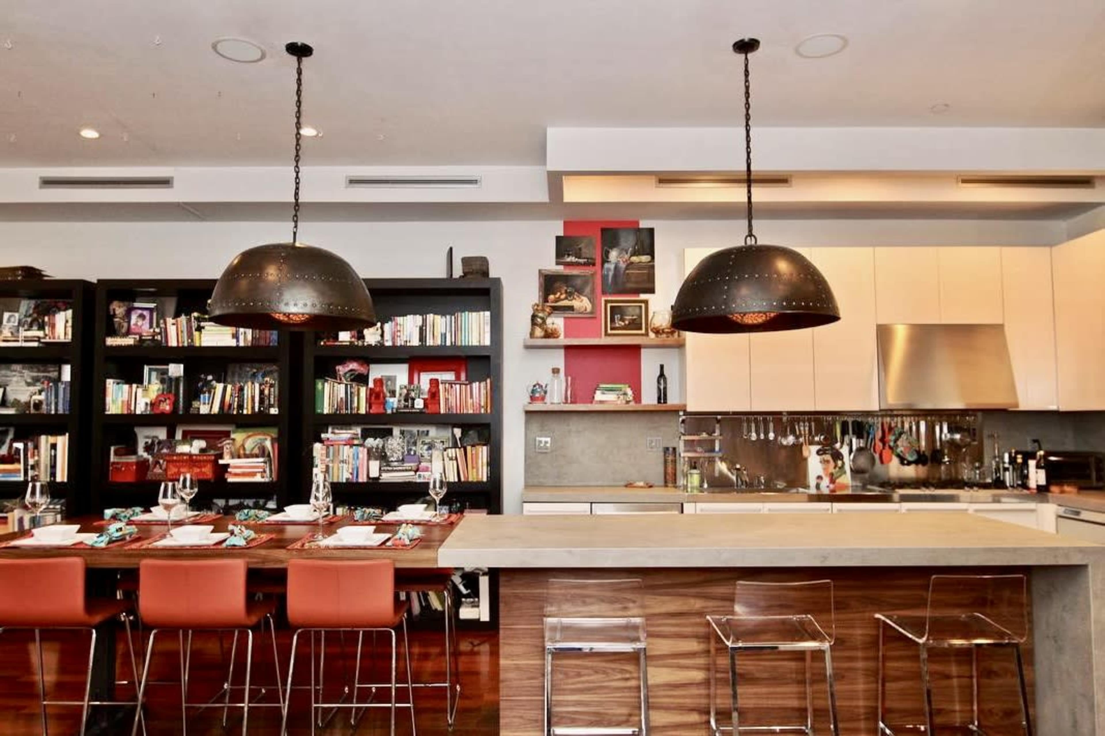 The image shows a modern kitchen with a large central island, two hanging pendant lights, and a dining area with a wooden table surrounded by orange chairs.