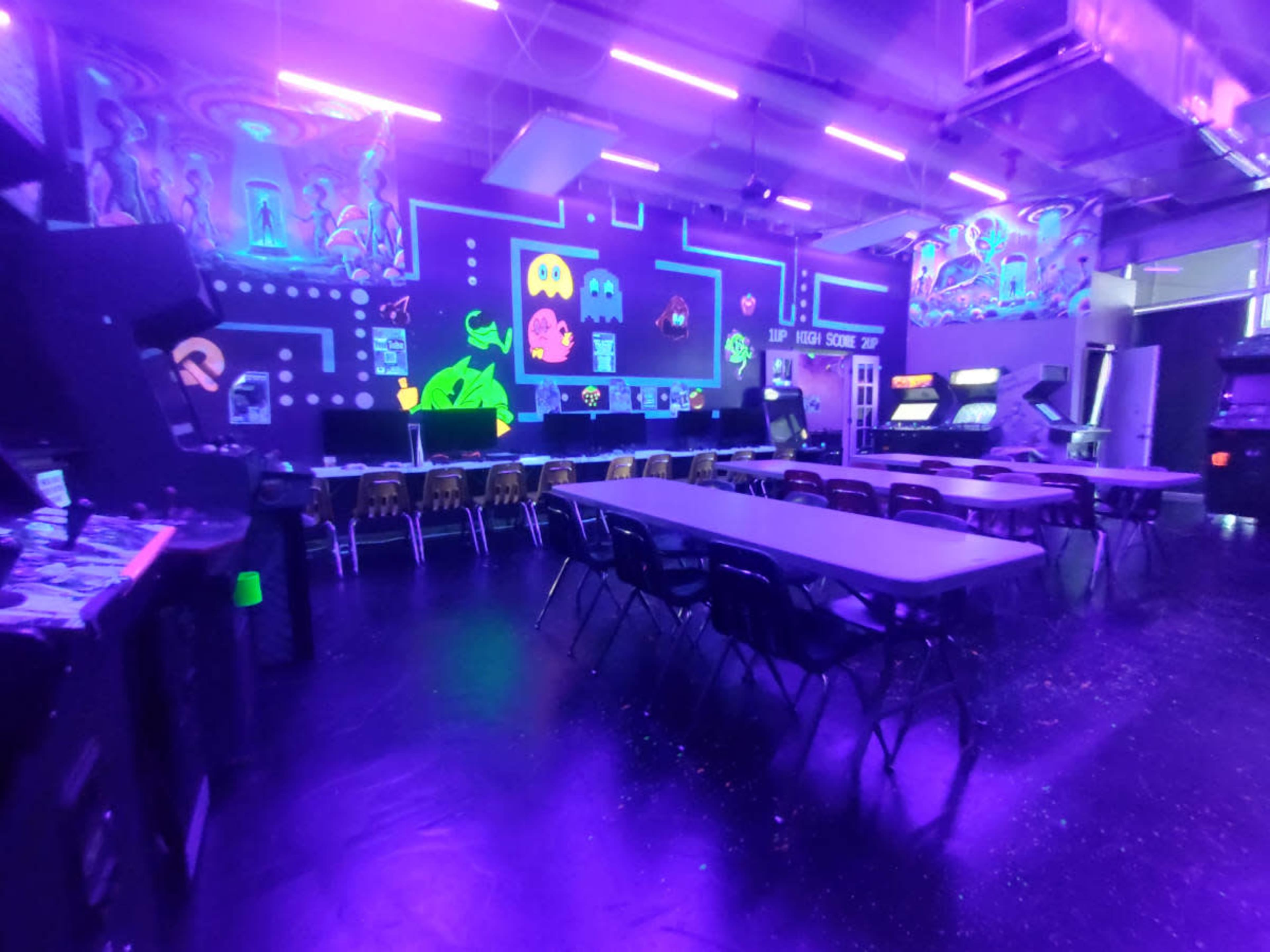The image shows an arcade room illuminated by purple lights, featuring various gaming machines and tables arranged in rows.