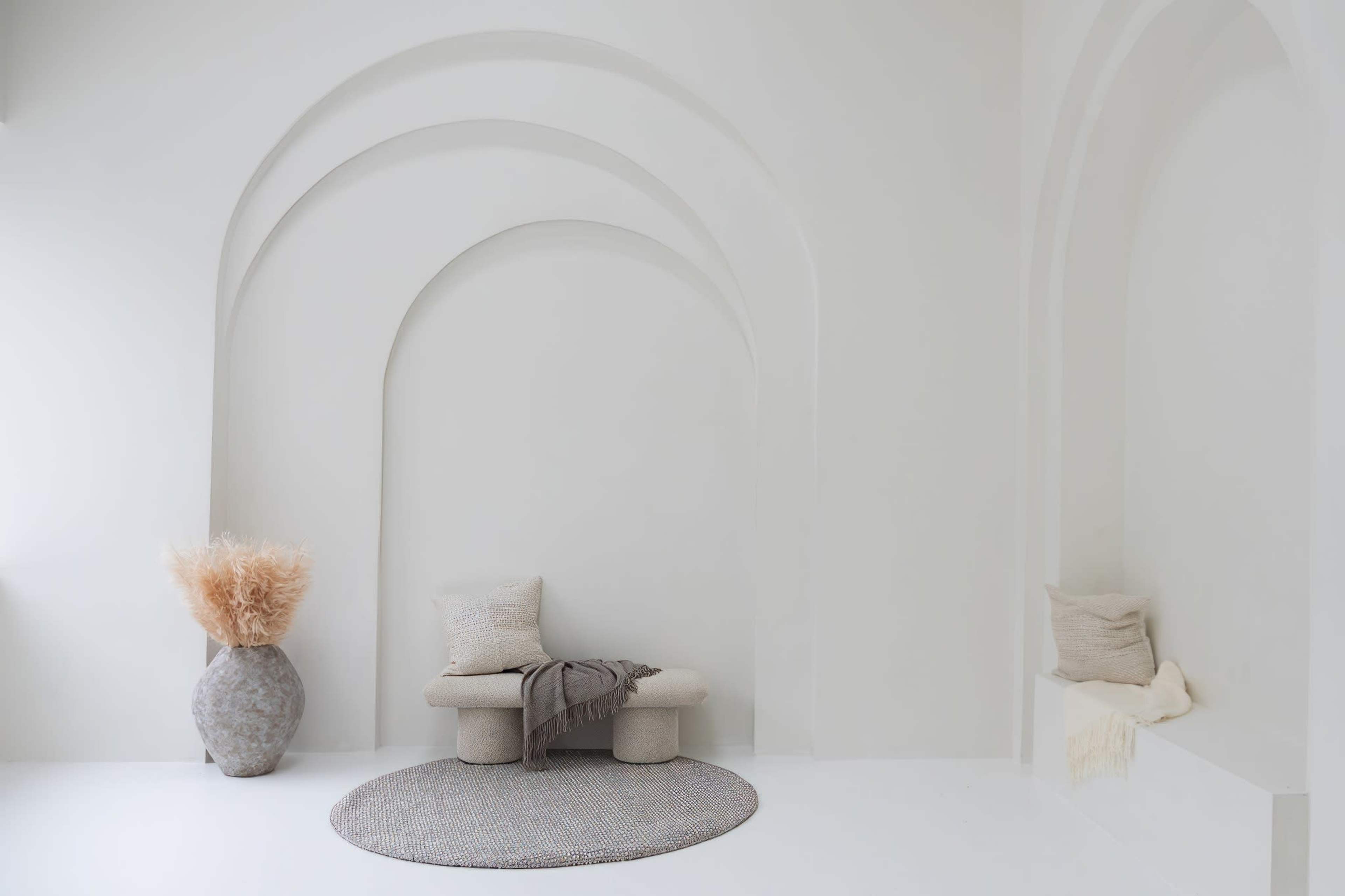 A minimalist interior space with arched wall recesses, a bench, decorative pillows, a round rug, and a large vase containing dried plants.