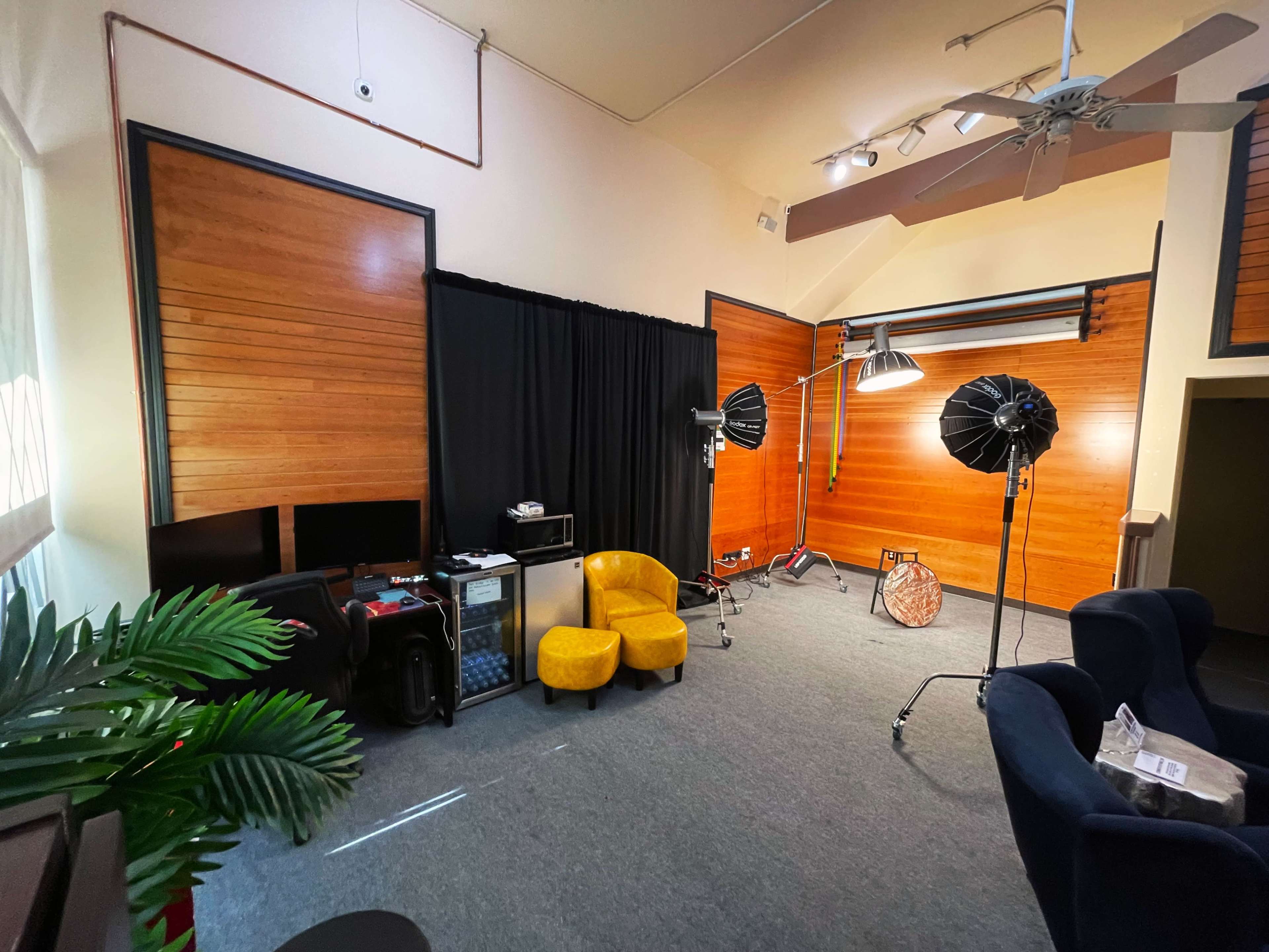 A photography studio featuring wooden walls, a black backdrop, two light stands with softboxes, and a pair of yellow armchairs.