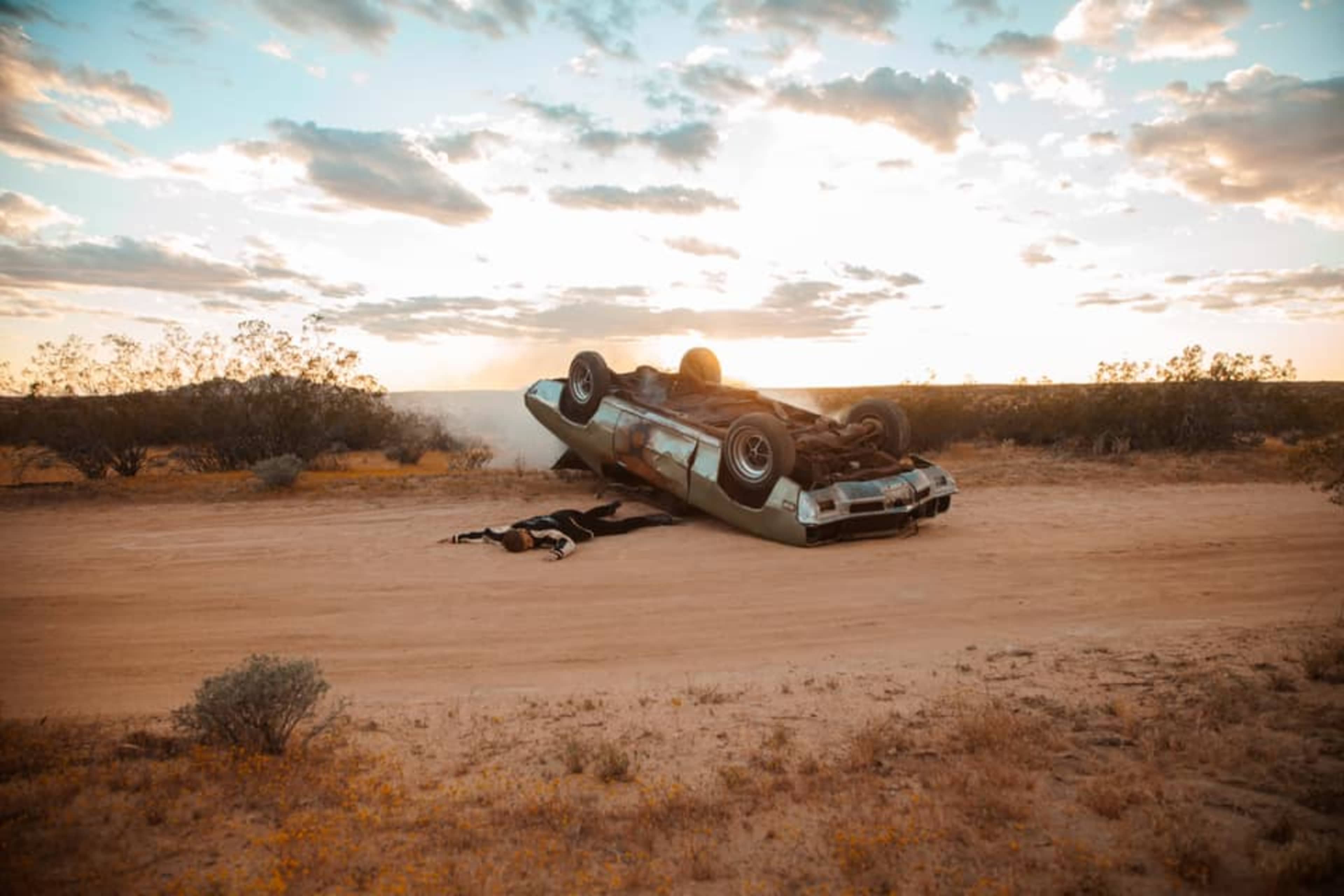 An overturned car lies on a dirt road in a desert landscape under a cloudy sky at sunset.